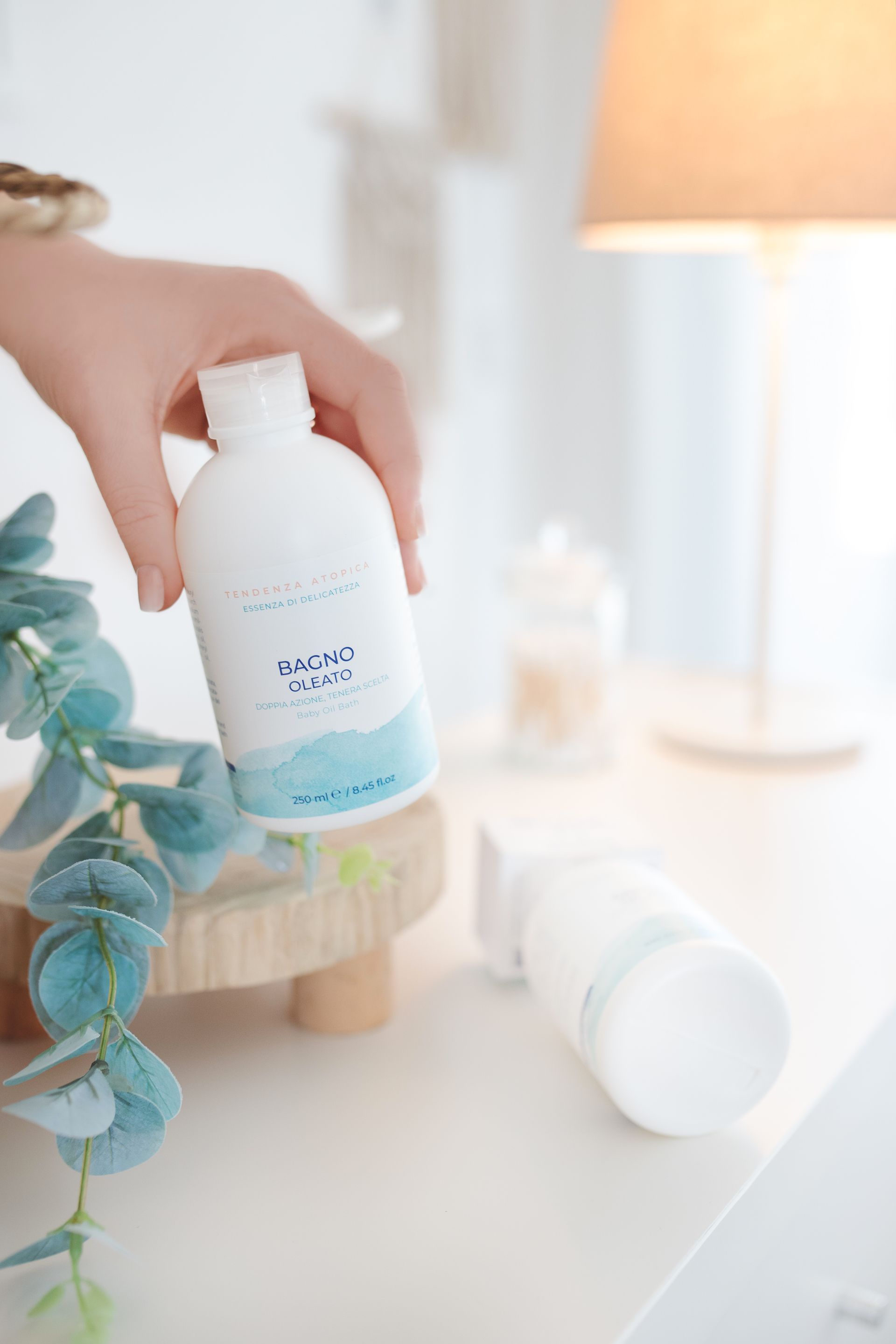 Mano che regge una bottiglia bianca con etichetta blu. Un'altra bottiglia è appoggiata accanto a essa su un tavolo bianco, accanto a un rametto di foglie blu.