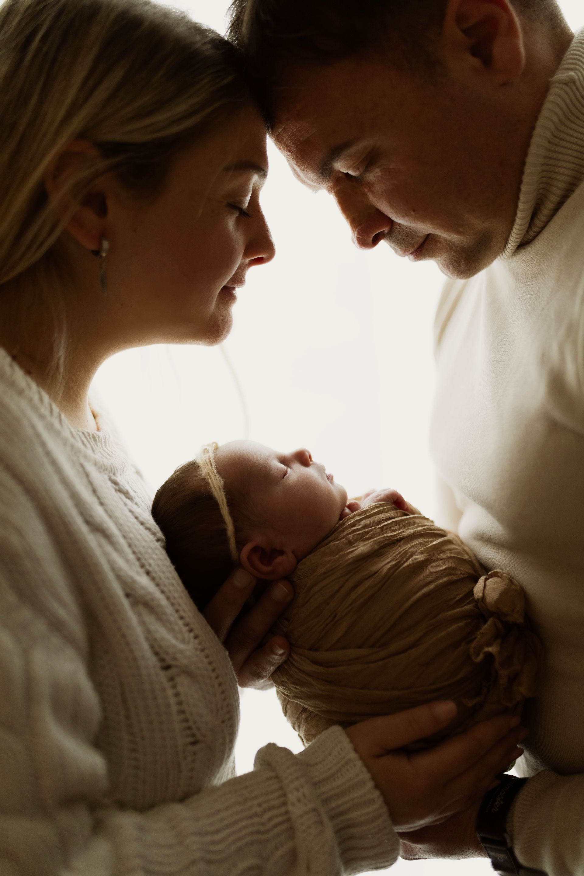 I genitori cullano dolcemente un neonato, toccandosi la fronte. Il bambino è avvolto in una coperta marrone; maglioncini color crema. Sfondo bianco e morbido.