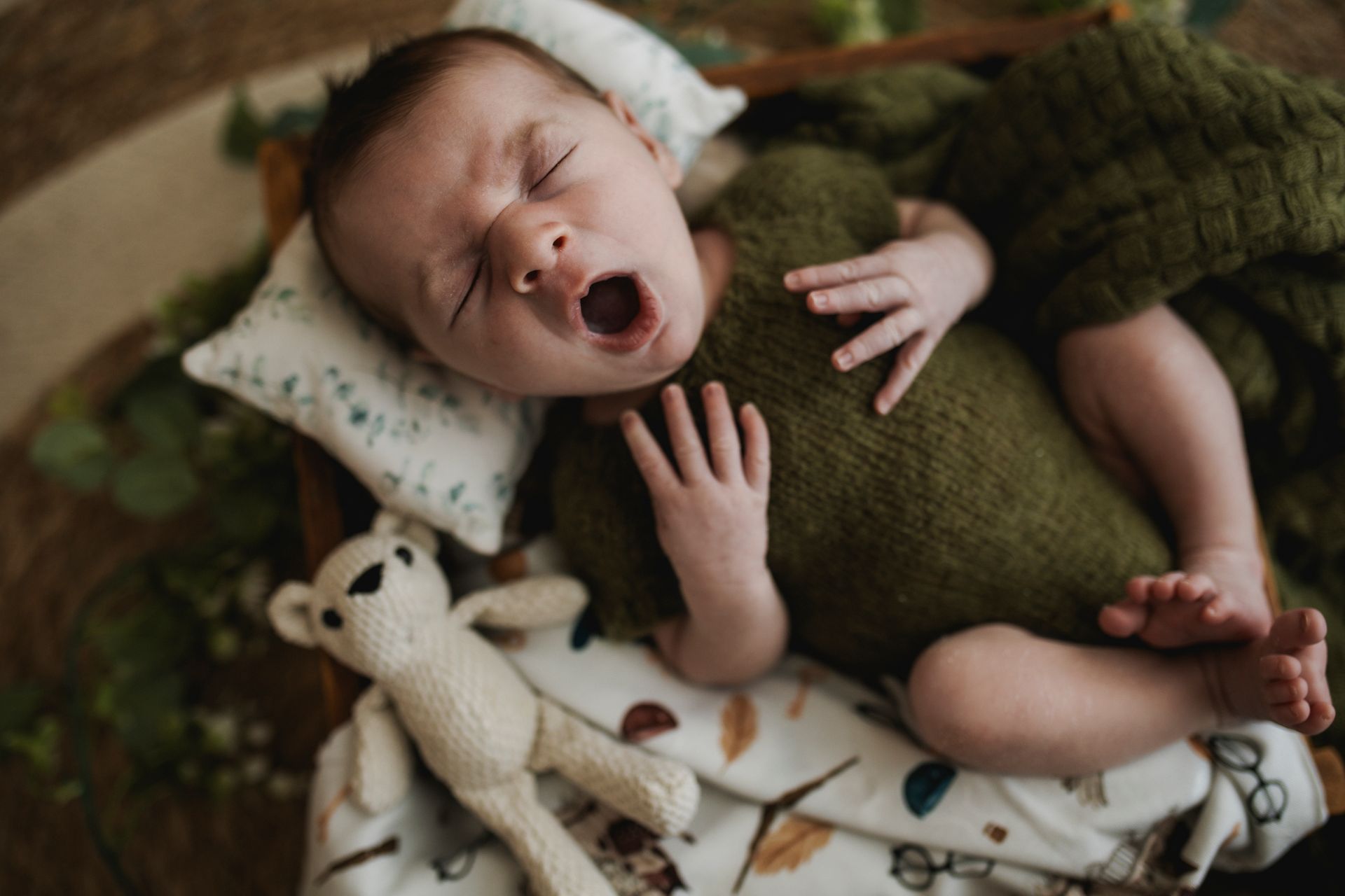 Un neonato che sbadiglia, indossa un vestito verde, rannicchiato in una cassa di legno con un orsacchiotto e una coperta.
