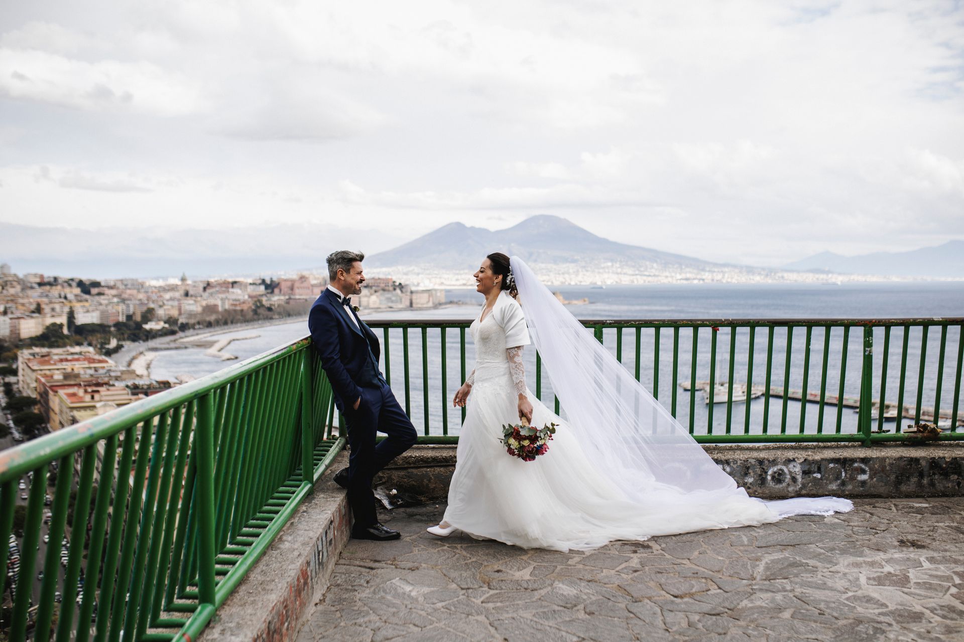 Sposi su un tetto con vista sulla città e su un vulcano. La sposa tiene in mano dei fiori, lo sposo è appoggiato a una ringhiera.