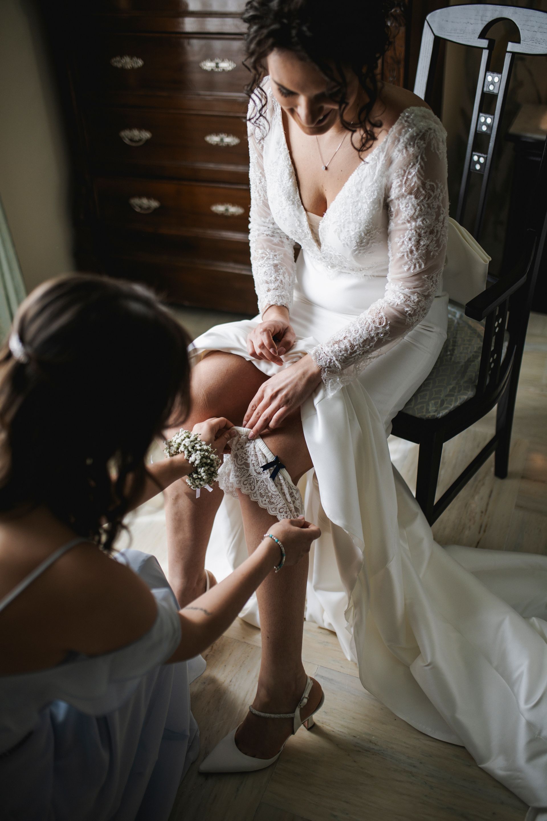 Sposa in abito da sposa, damigella che le sistema la giarrettiera sulla gamba. Interno, luce naturale.