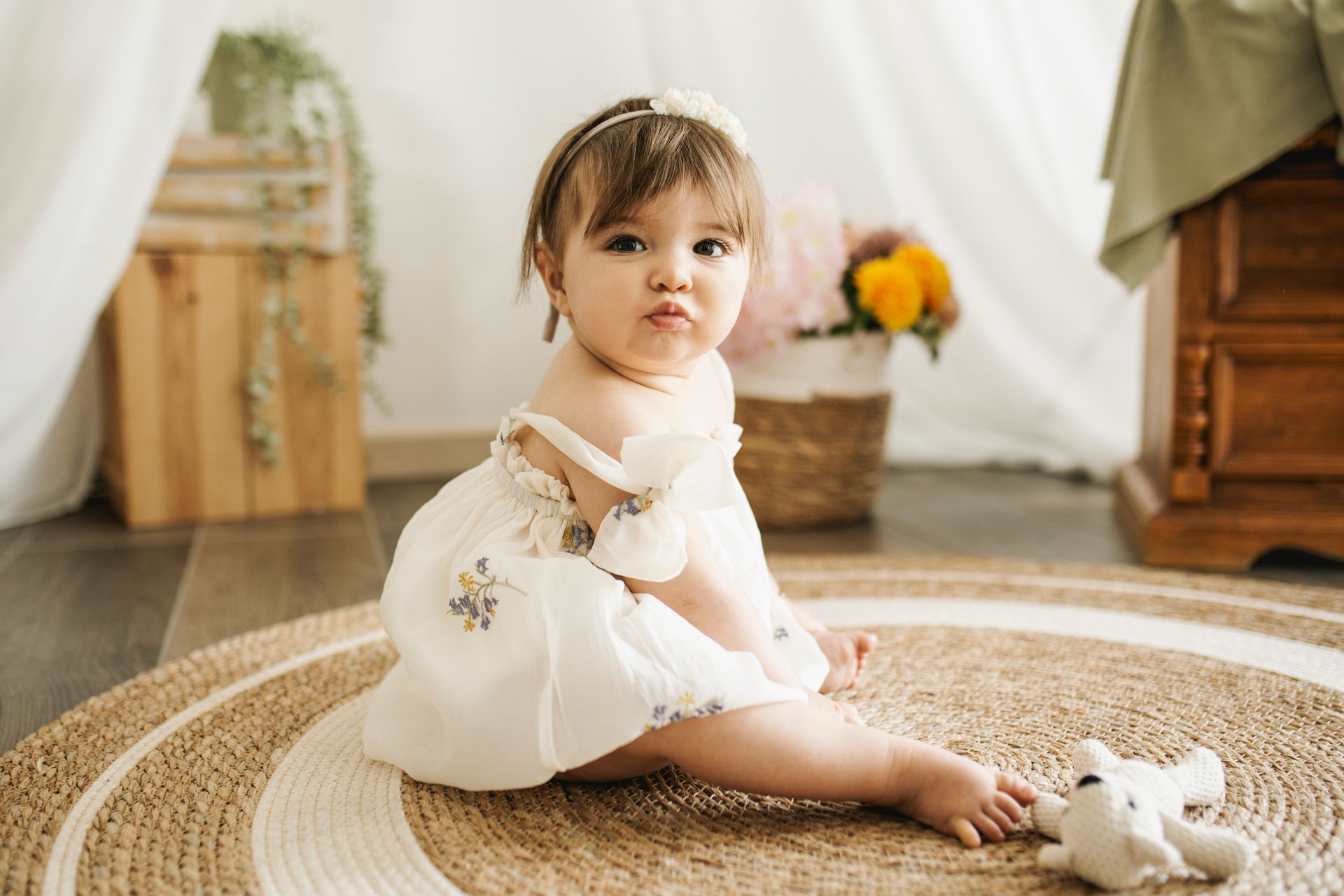 Una bambina seduta su un tappeto rotondo, con indosso un abito bianco, guarda verso la telecamera, con fiori e legno sullo sfondo.