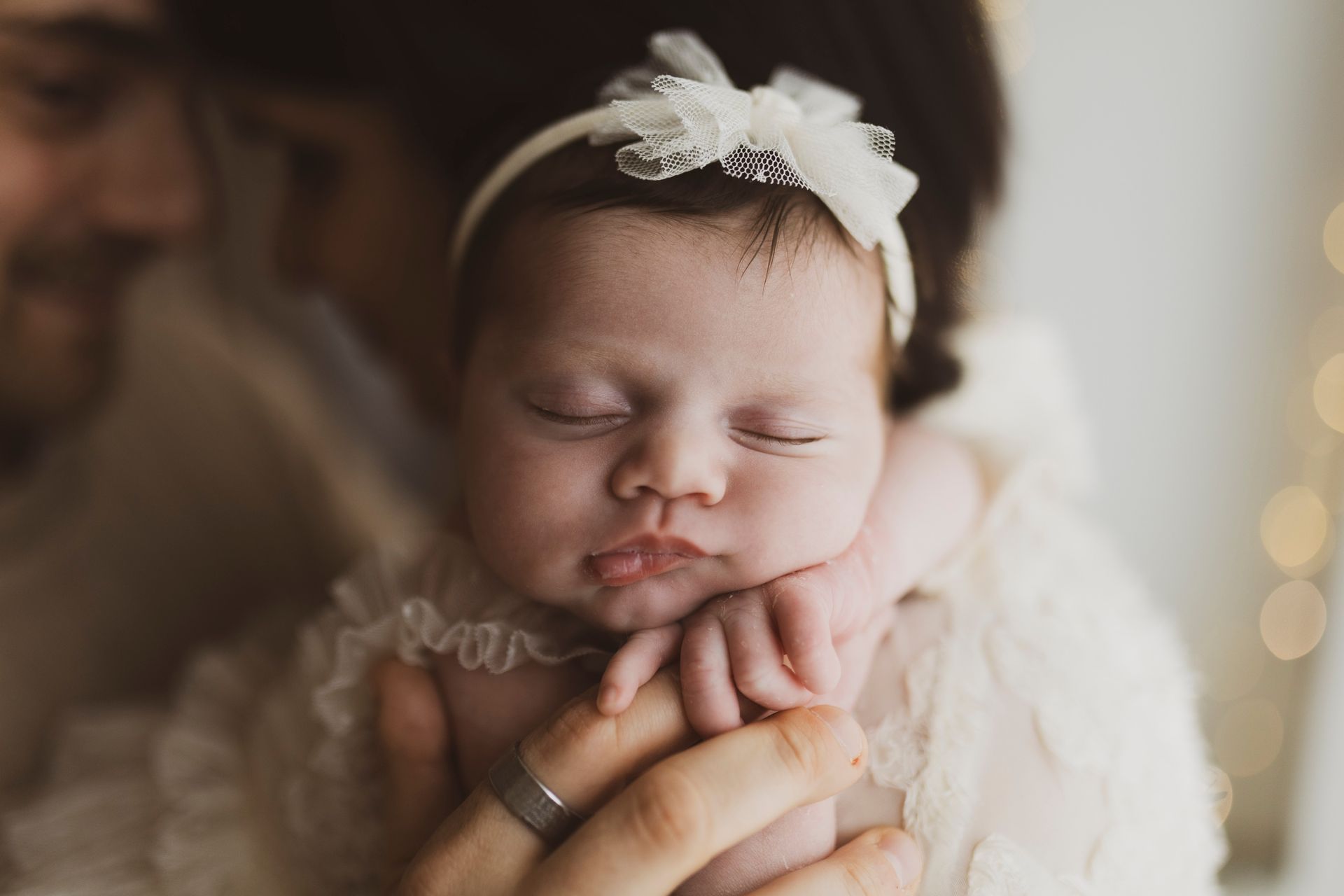 Neonato che dorme, tenuto in braccio dai genitori; indossa una fascia di pizzo e un vestitino.