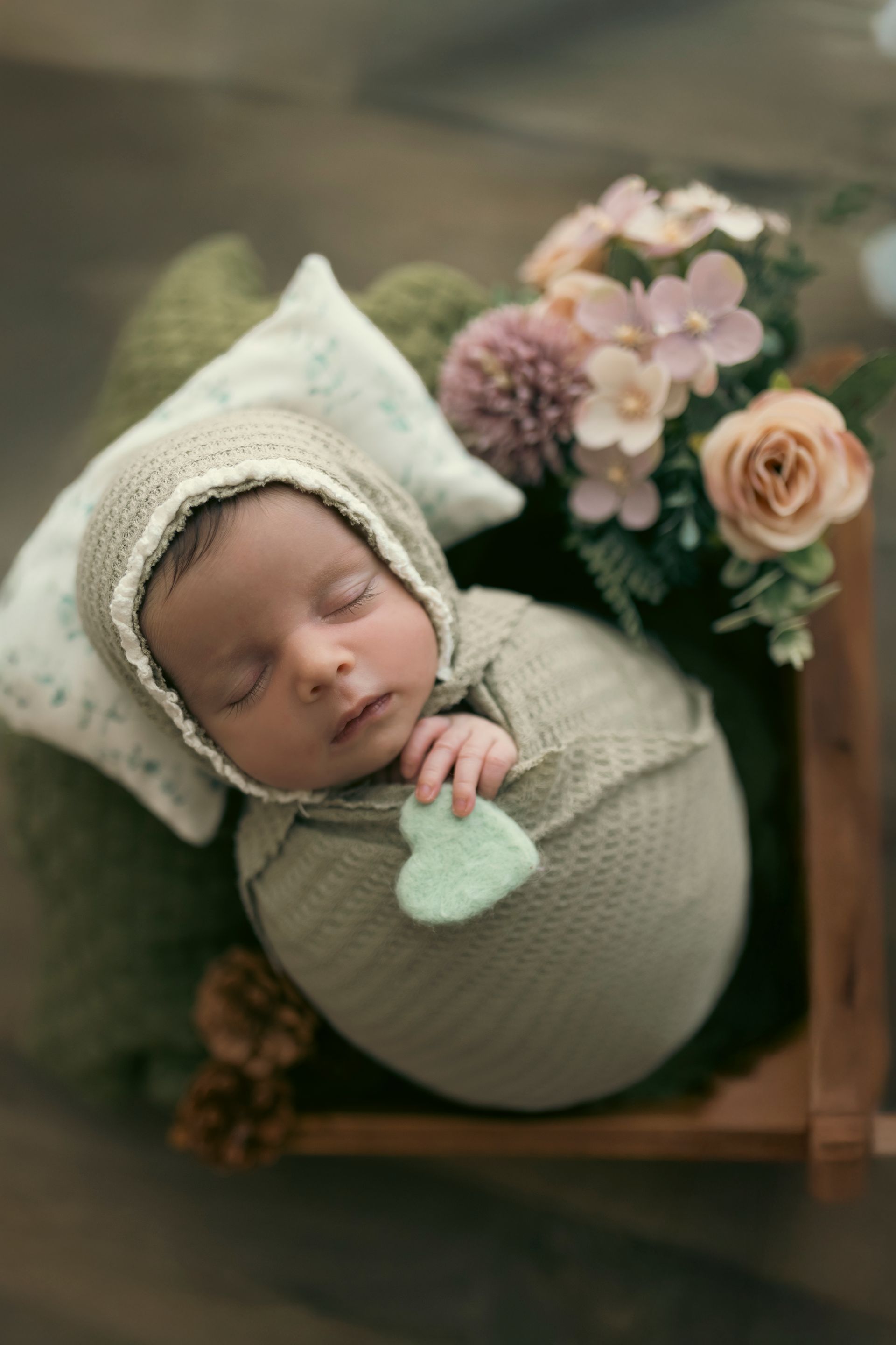 Neonato avvolto in fasce verdi, con indosso una cuffia, che dorme in una scatola di legno con dei fiori.