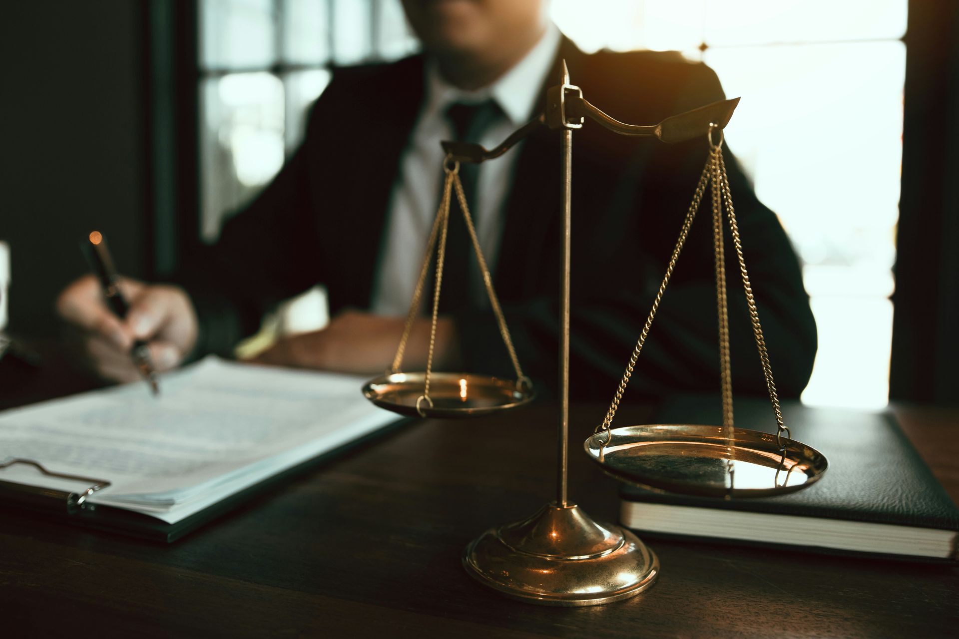 Scales of justice in focus, a blurred figure of a person in a suit writes at a desk.