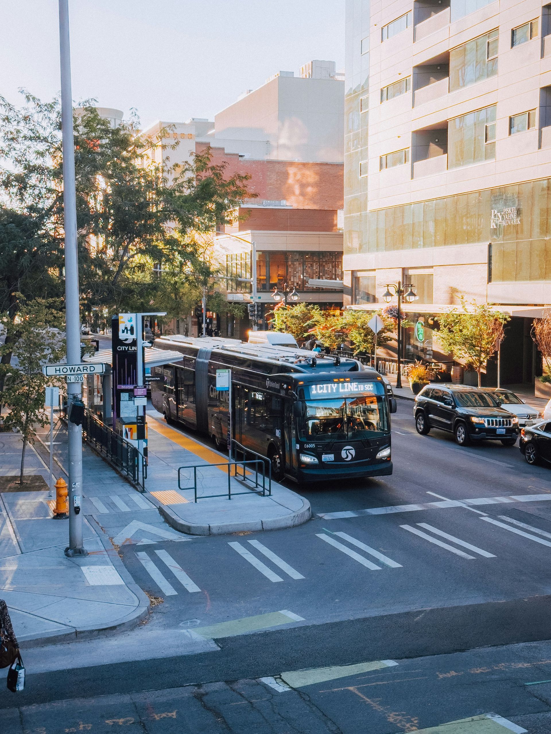 Bus at a stop in a city. Buildings, trees, and cars are visible. Sunny day.
