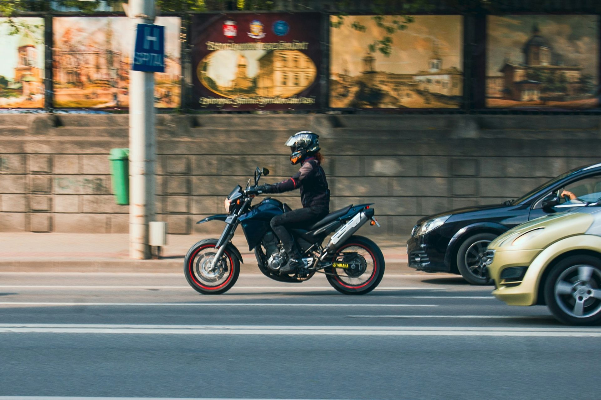 Motorcyclist rides a blue motorcycle on a city street, near cars and a wall with paintings.