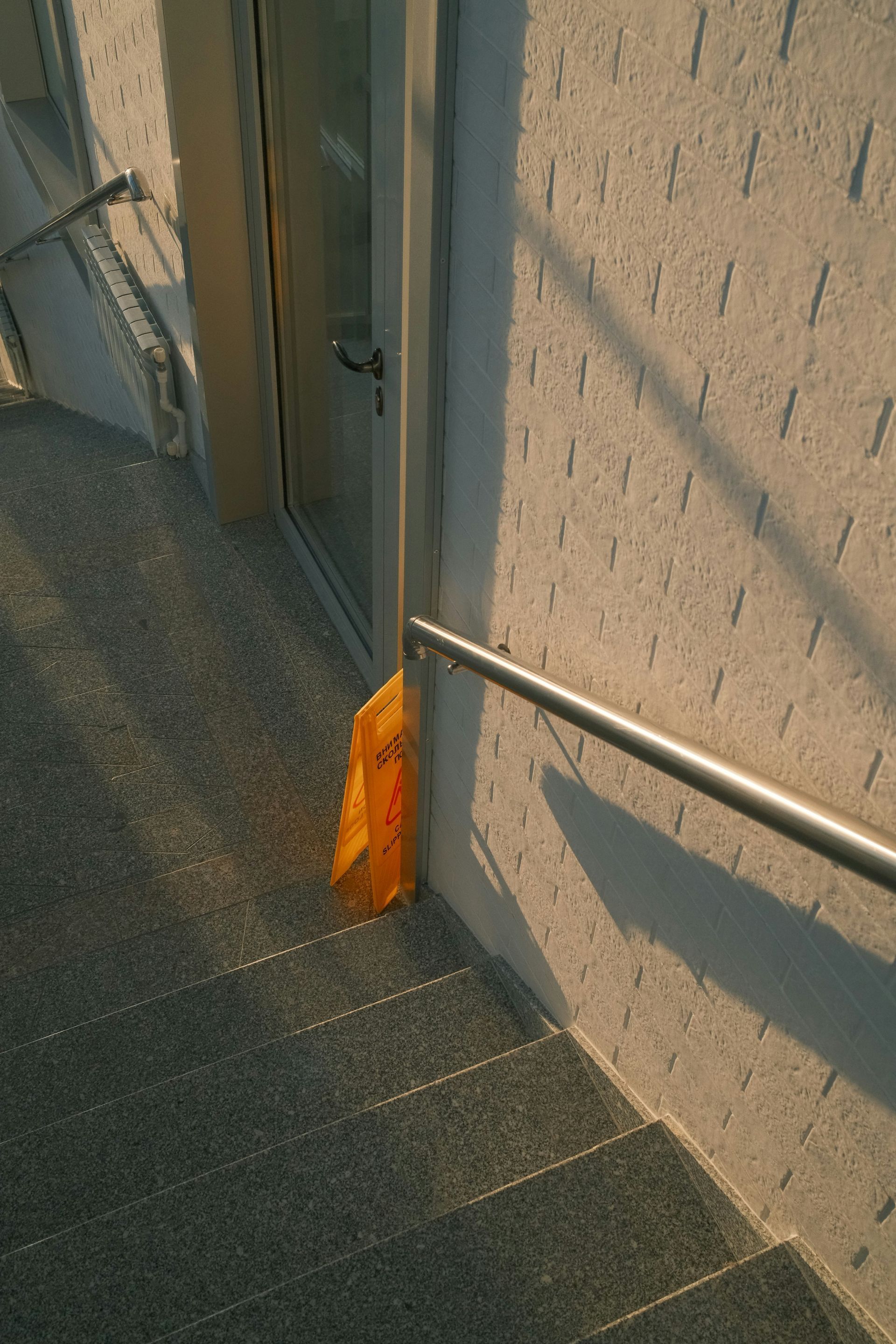 Yellow caution sign near a doorway and stainless steel handrail, placed at the top of outdoor stairs.