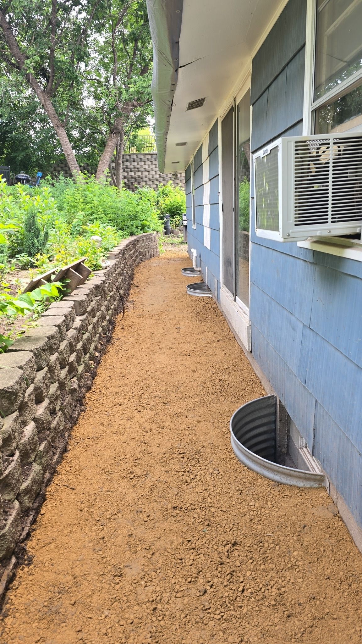 A narrow gravel path alongside a blue house with a window AC unit and retaining wall.