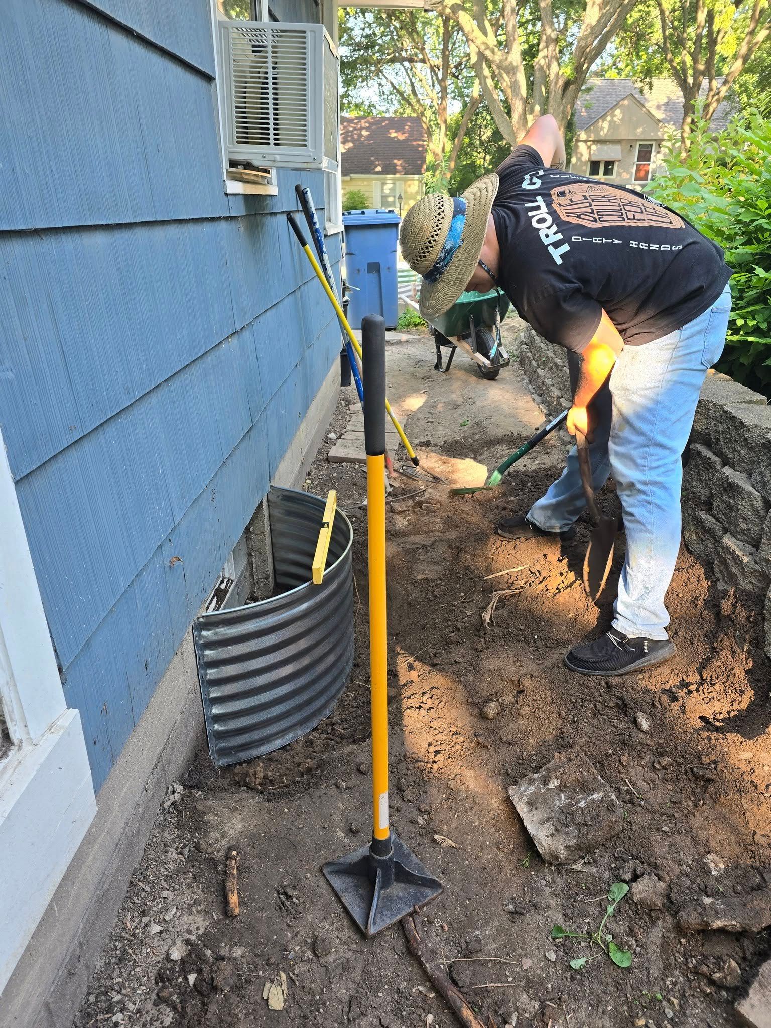 Person digging soil near a blue house; a window well is visible. A tool and dog are nearby.