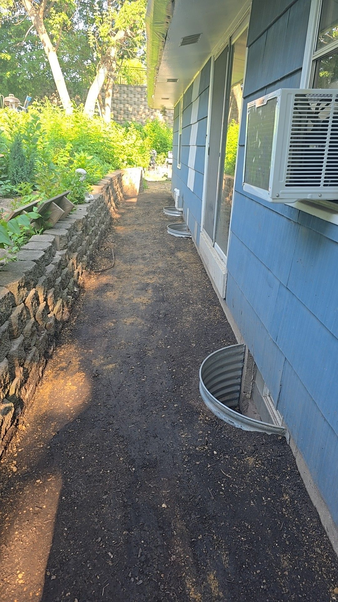 Long, narrow dirt path between a blue house and a stone retaining wall, with a window air conditioner.