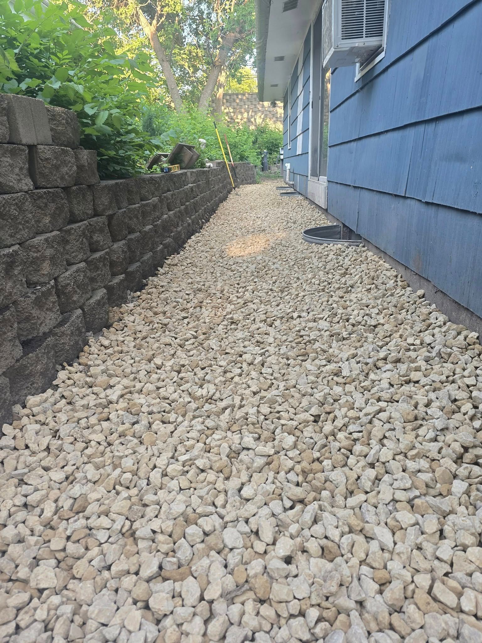 Gravel path between a blue house and a retaining wall of stacked dark gray blocks, with greenery visible above.