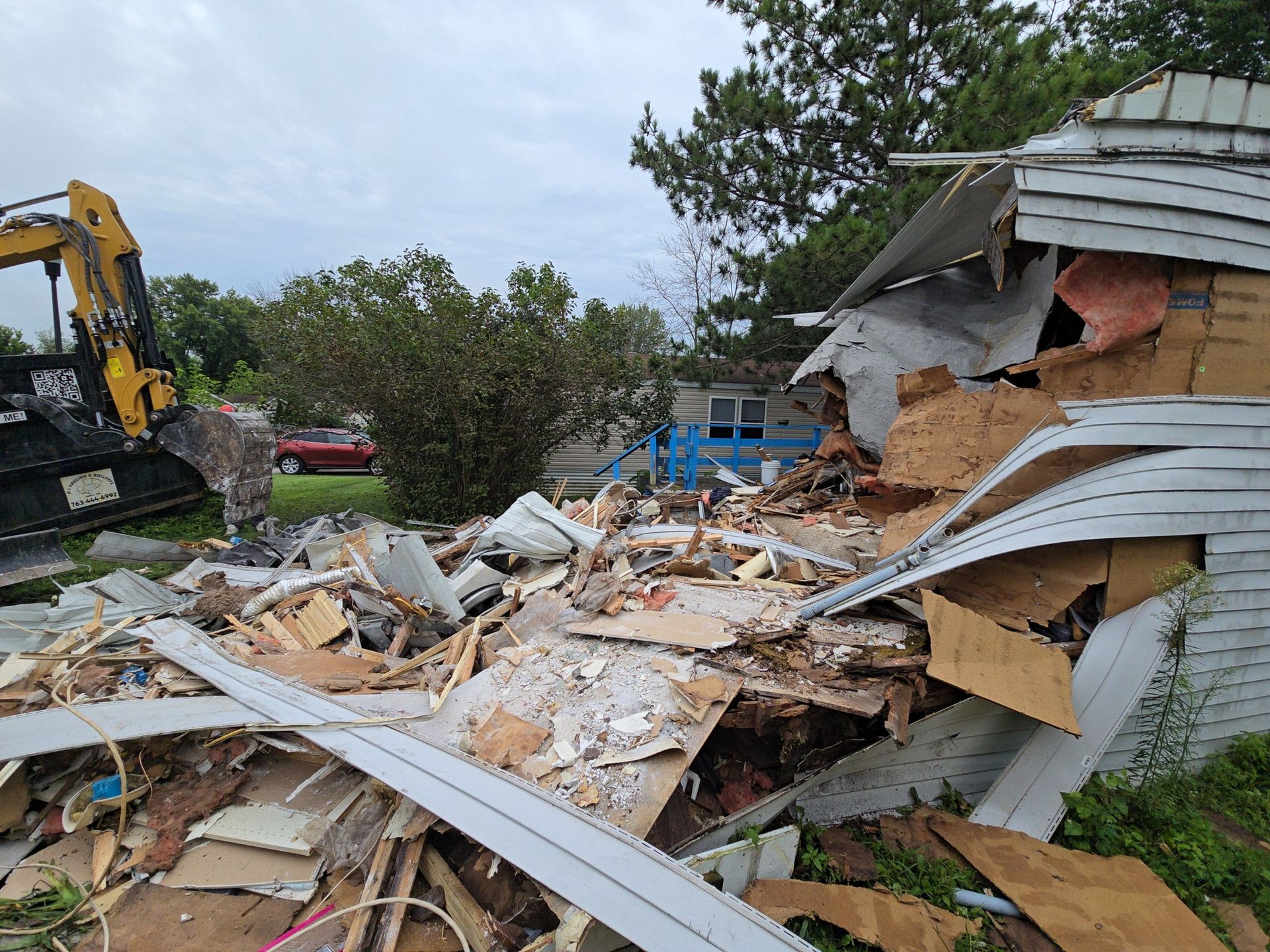 Demolished house with a backhoe. Debris pile in front; cloudy sky.