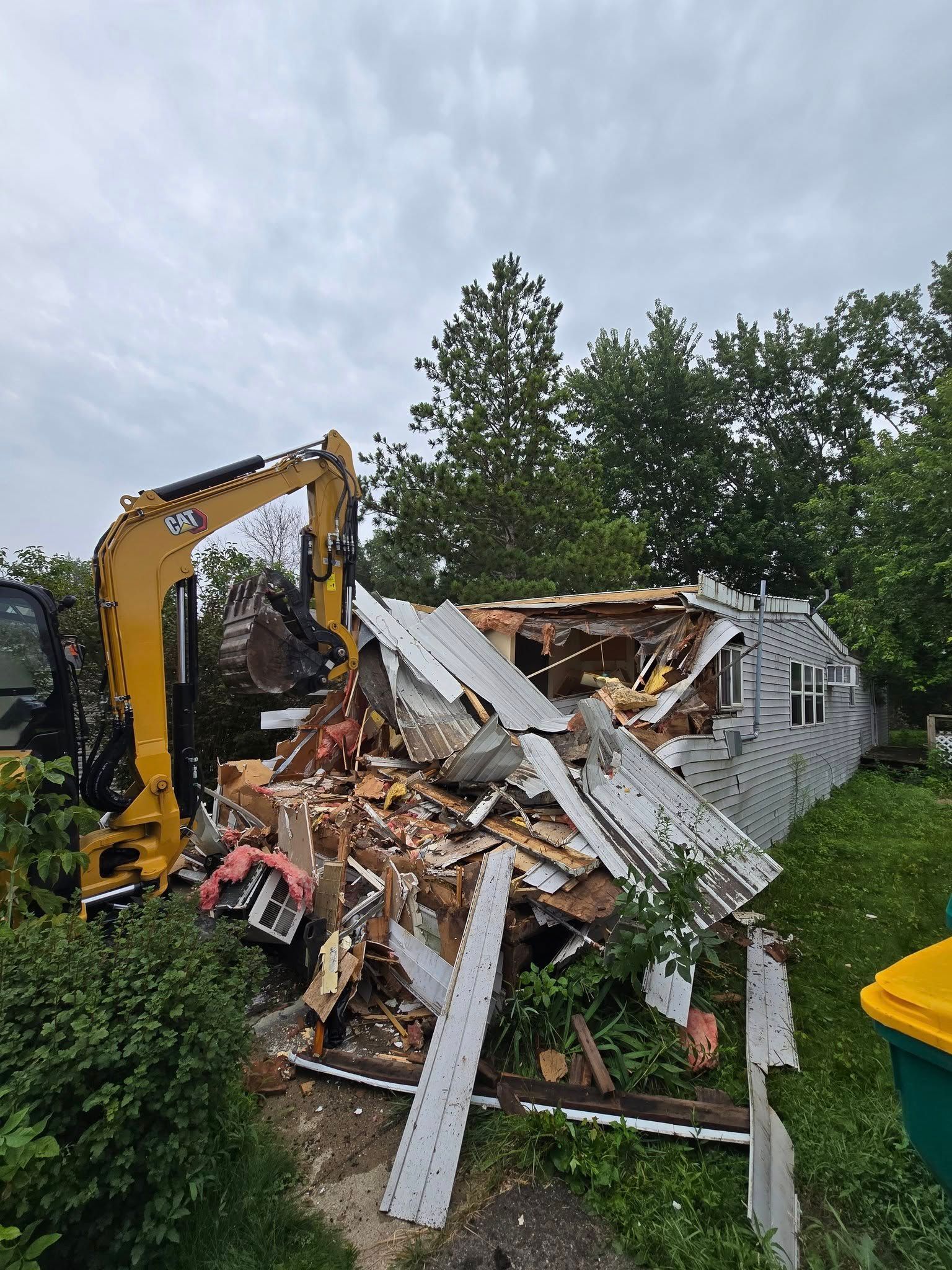 A yellow excavator demolishes a house. Debris and exposed structure visible. Overcast sky and green foliage.