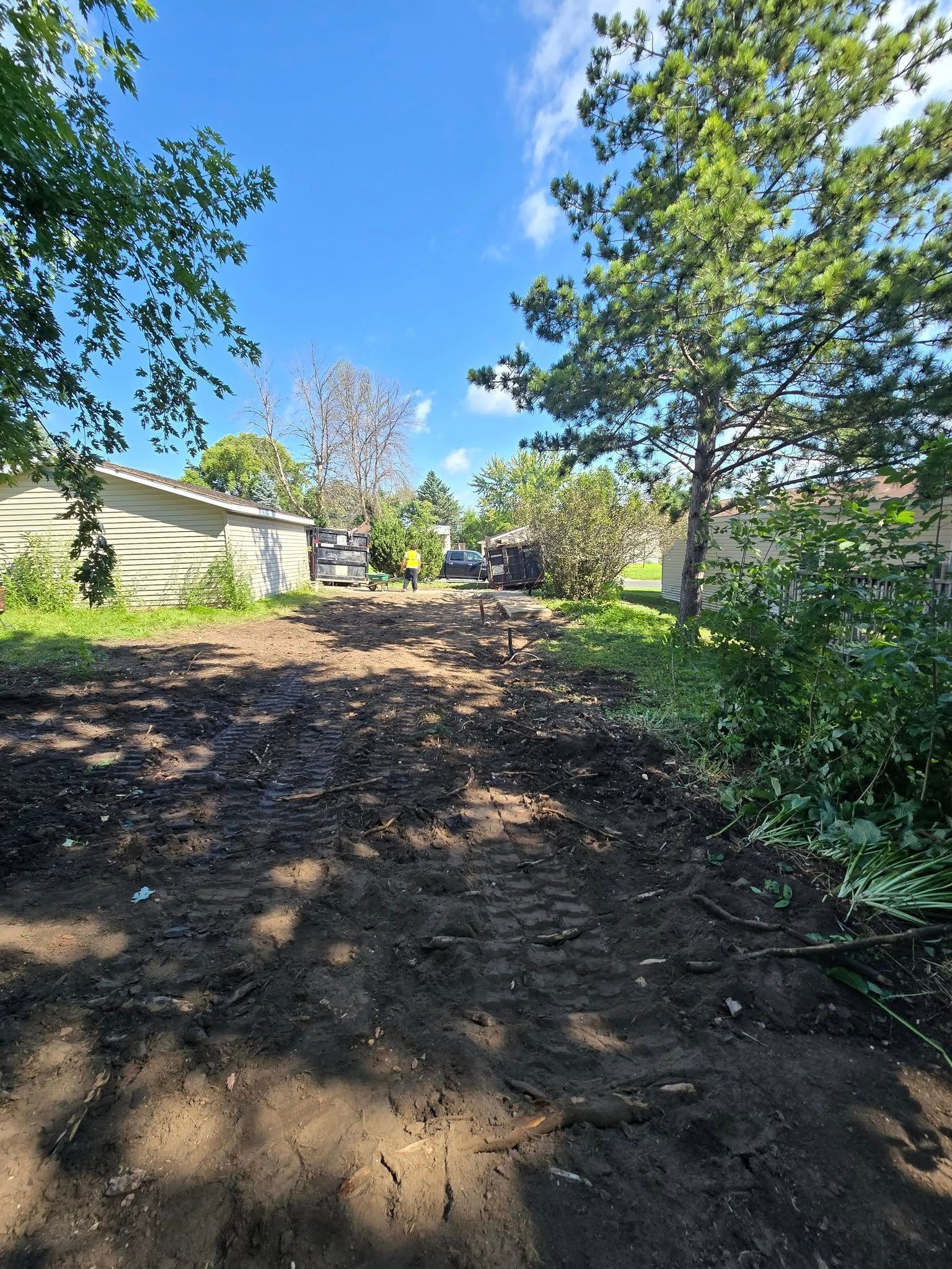 Dirt path with construction vehicles, bordered by trees and buildings, under a blue sky.