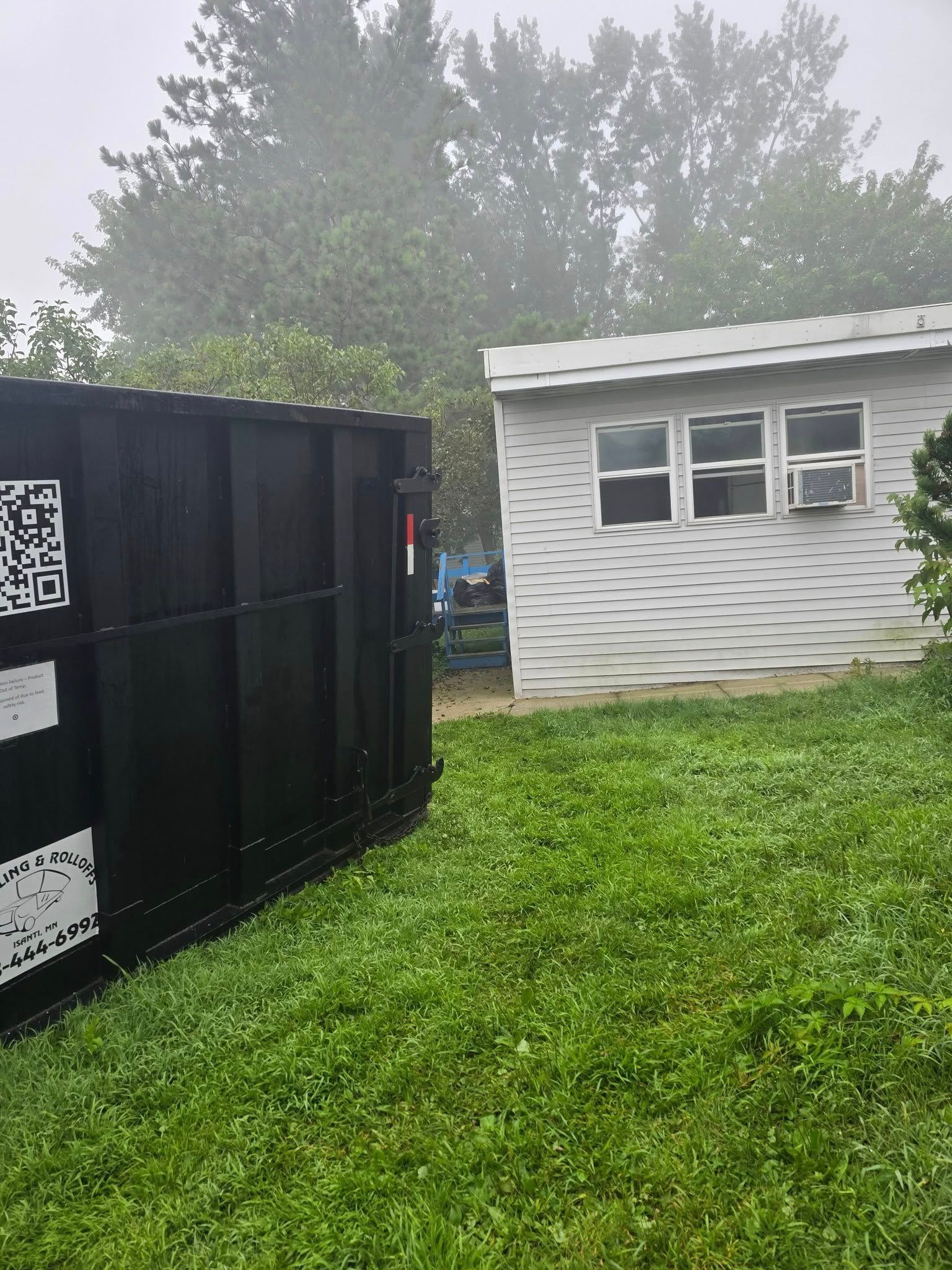 Black dumpster on green lawn, next to a small white building with windows; foggy background.