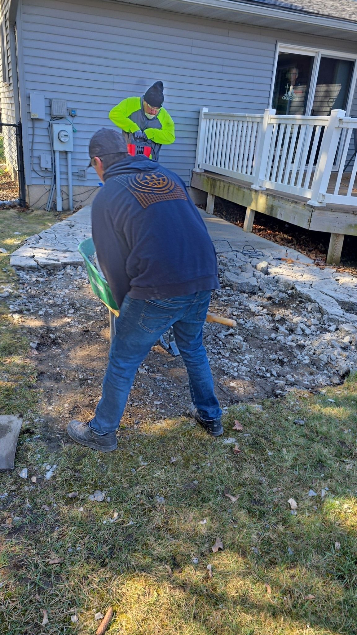 Two workers outdoors; one shovels debris, the other stands nearby. House and deck in background.