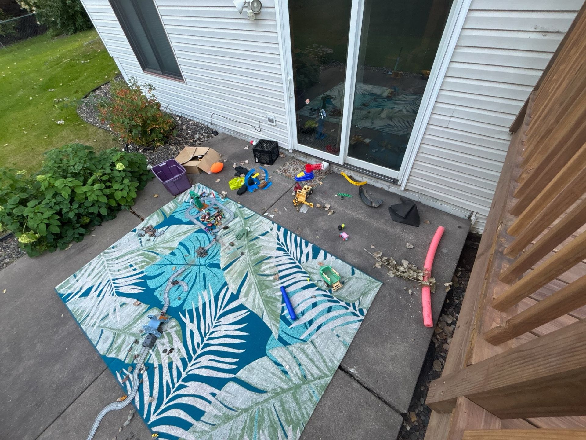 Patio with rug and scattered toys in front of a sliding glass door. A pool noodle sits nearby.