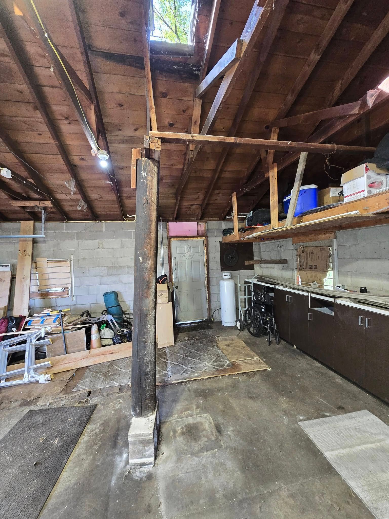 Inside of a cluttered garage: a support beam, wooden roof structure, workbenches, and open door.