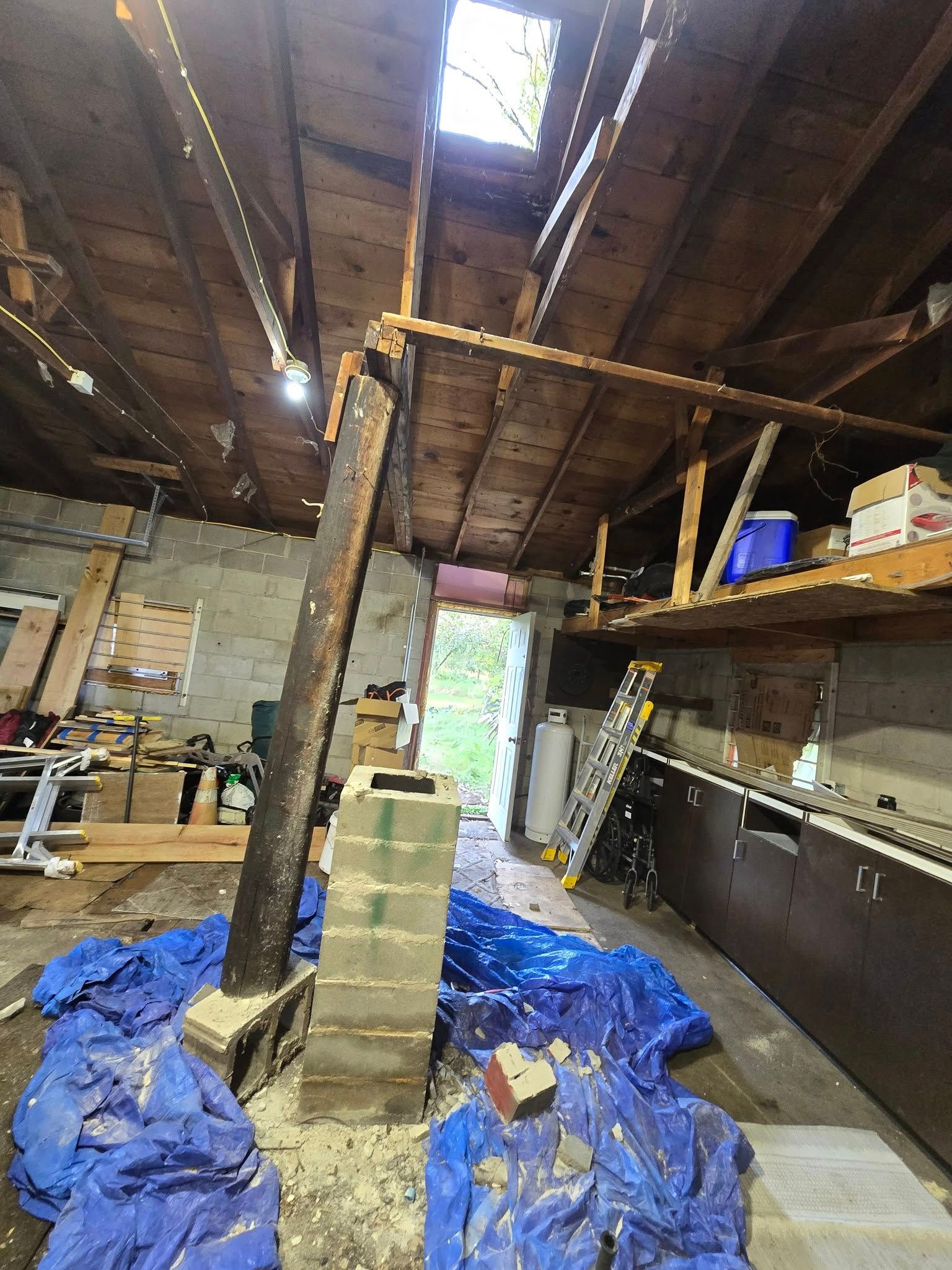 Interior of a cluttered workshop; support beam, exposed rafters, skylight, shelves with boxes, tarp on the floor.