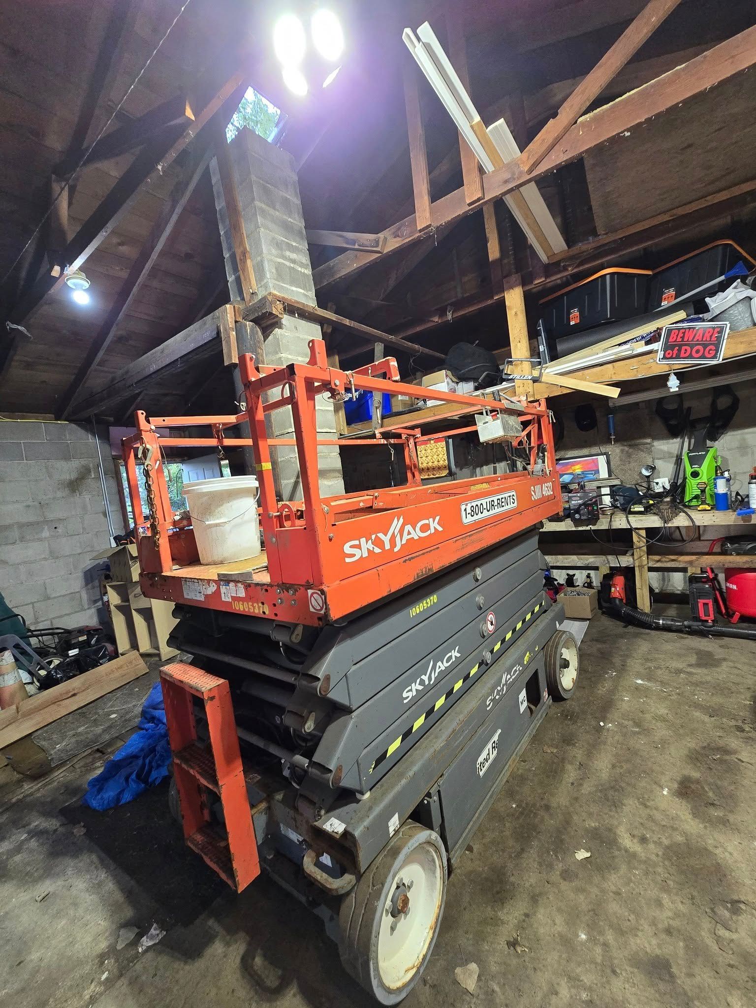 A scissor lift in a cluttered workshop with exposed wooden beams, an overhead light, and tools.