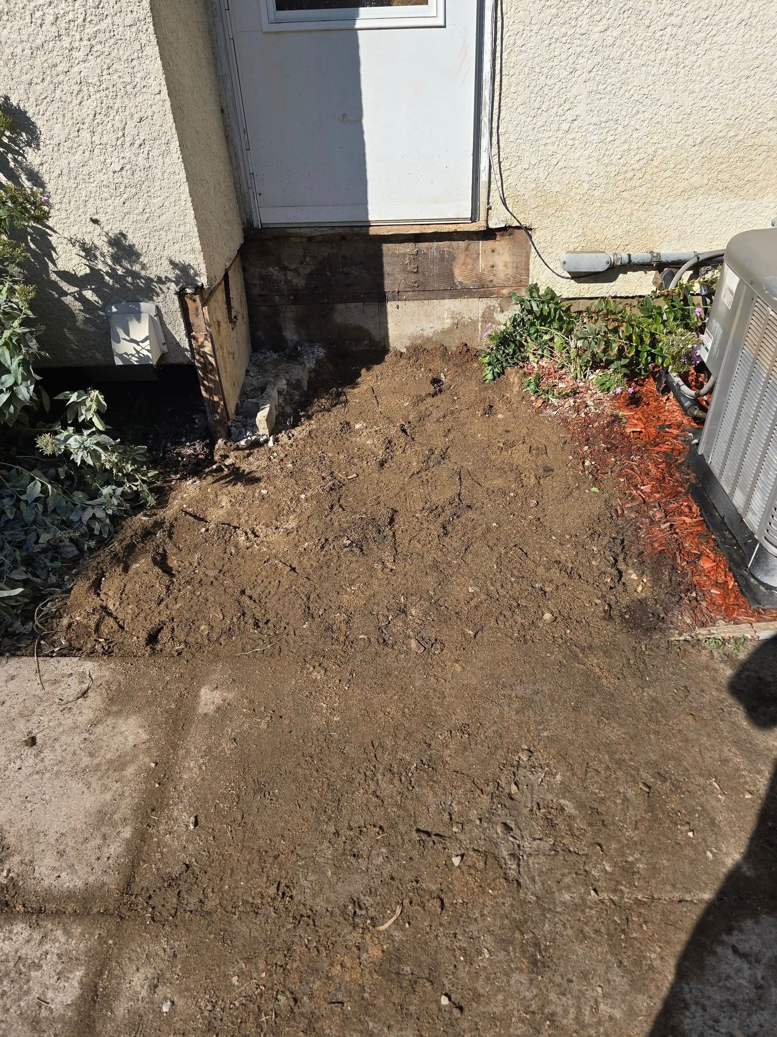 Dirt pile in front of a white door with a concrete slab; side of the building is textured beige.