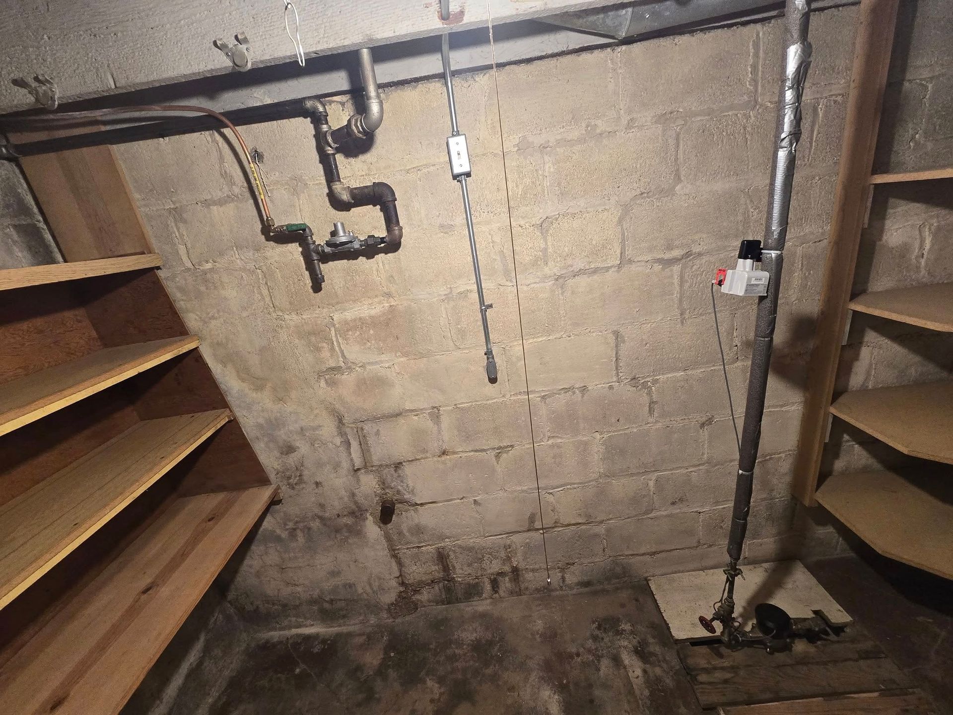 Basement corner with concrete block wall, shelves, pipes, and electrical conduit.