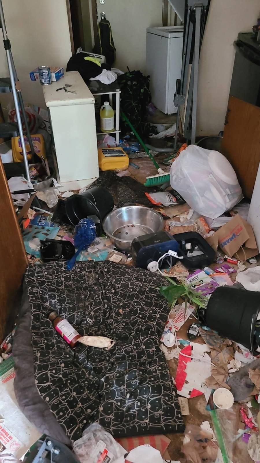 Room filled with trash, debris, and scattered items. White refrigerator, washer, and bags of trash are visible.