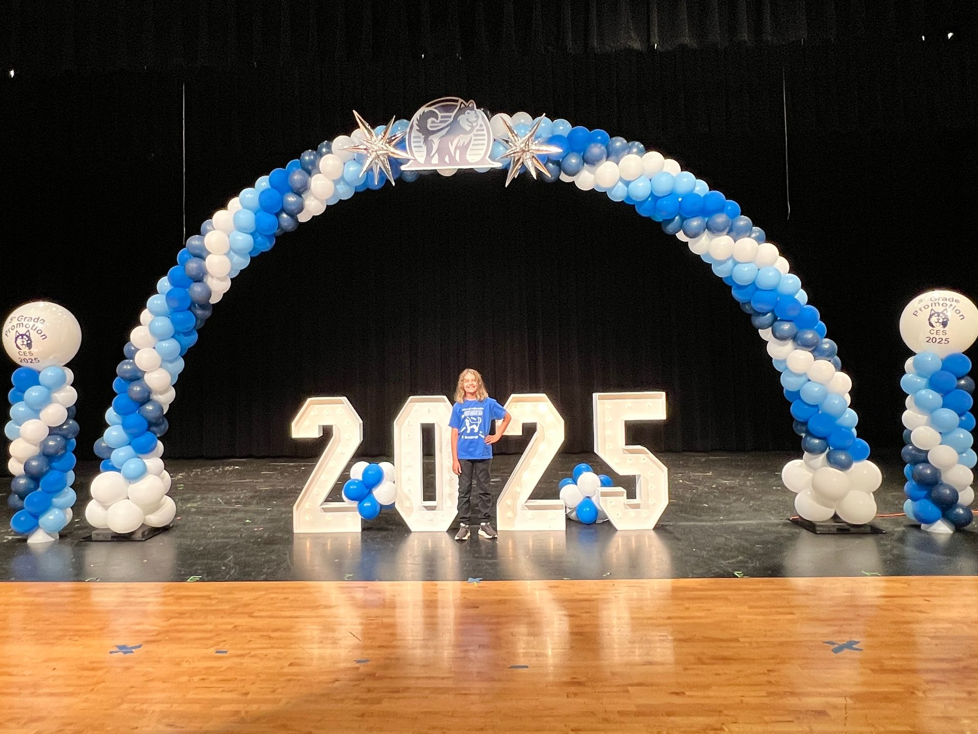 Young person stands before 2025 display on a stage, blue and white balloon arch overhead. One balloon column on each side of stage.