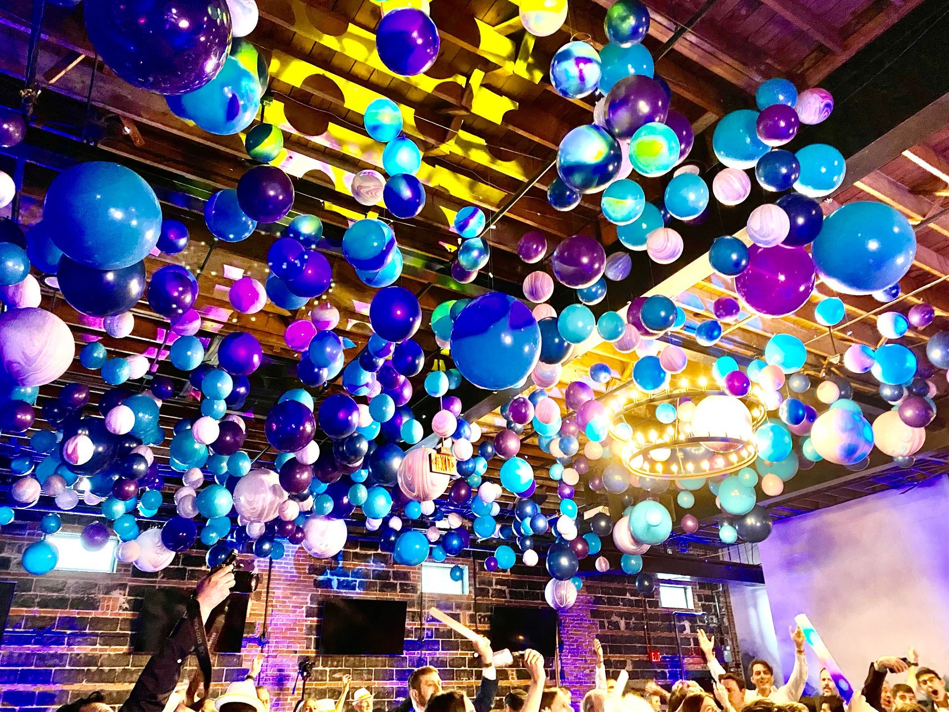 A crowd of people are standing in a room with balloons hanging from the ceiling.