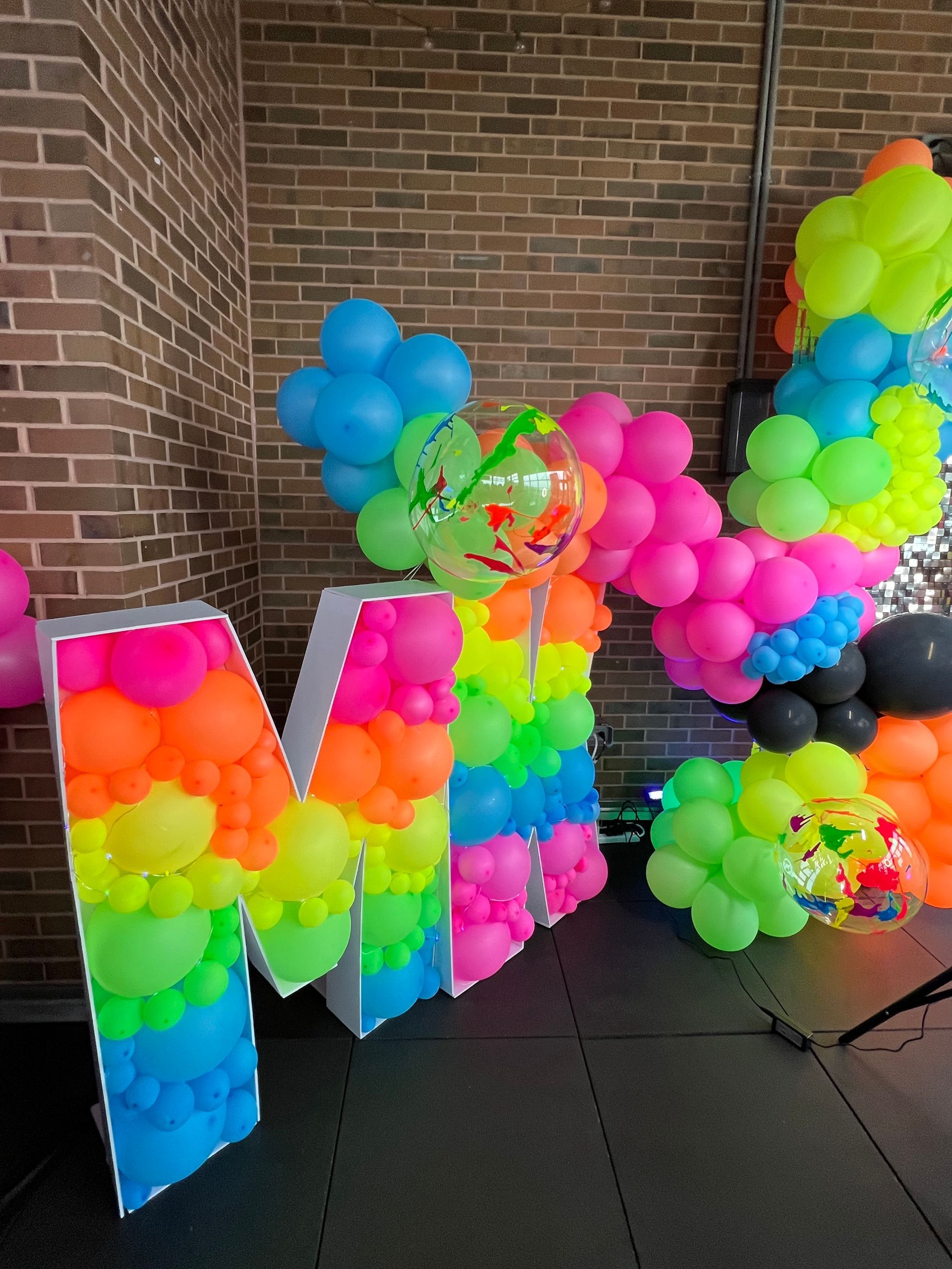 The letter m is made of neon balloons in front of a brick wall