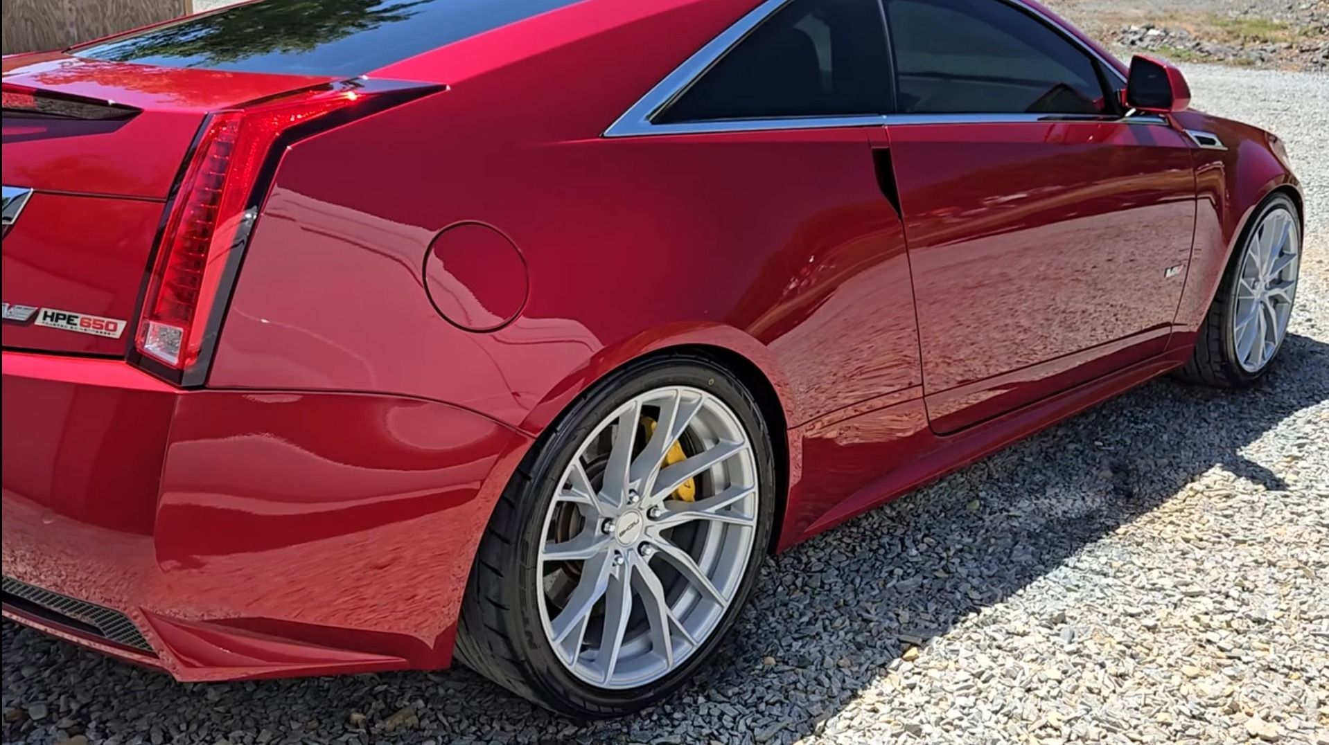 Red Cadillac coupe with silver wheels on gravel.