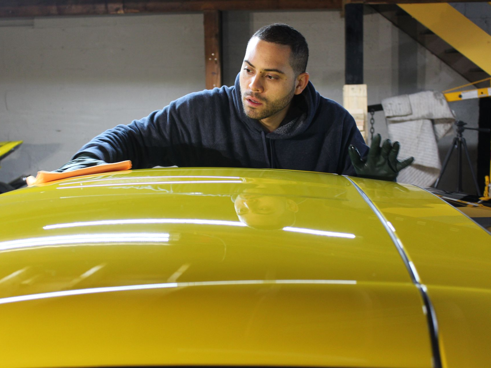 Man in blue hoodie polishing a bright yellow car roof in a garage.