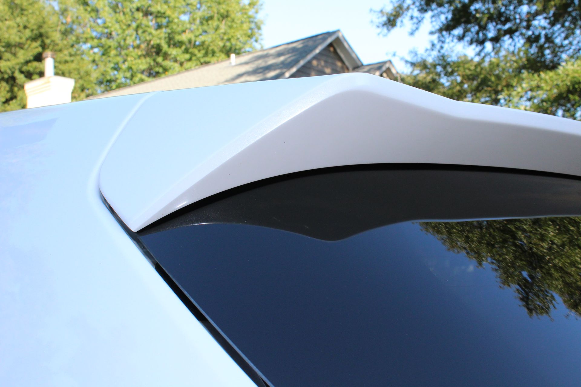 White car roof with a spoiler, against a background of trees and a house.