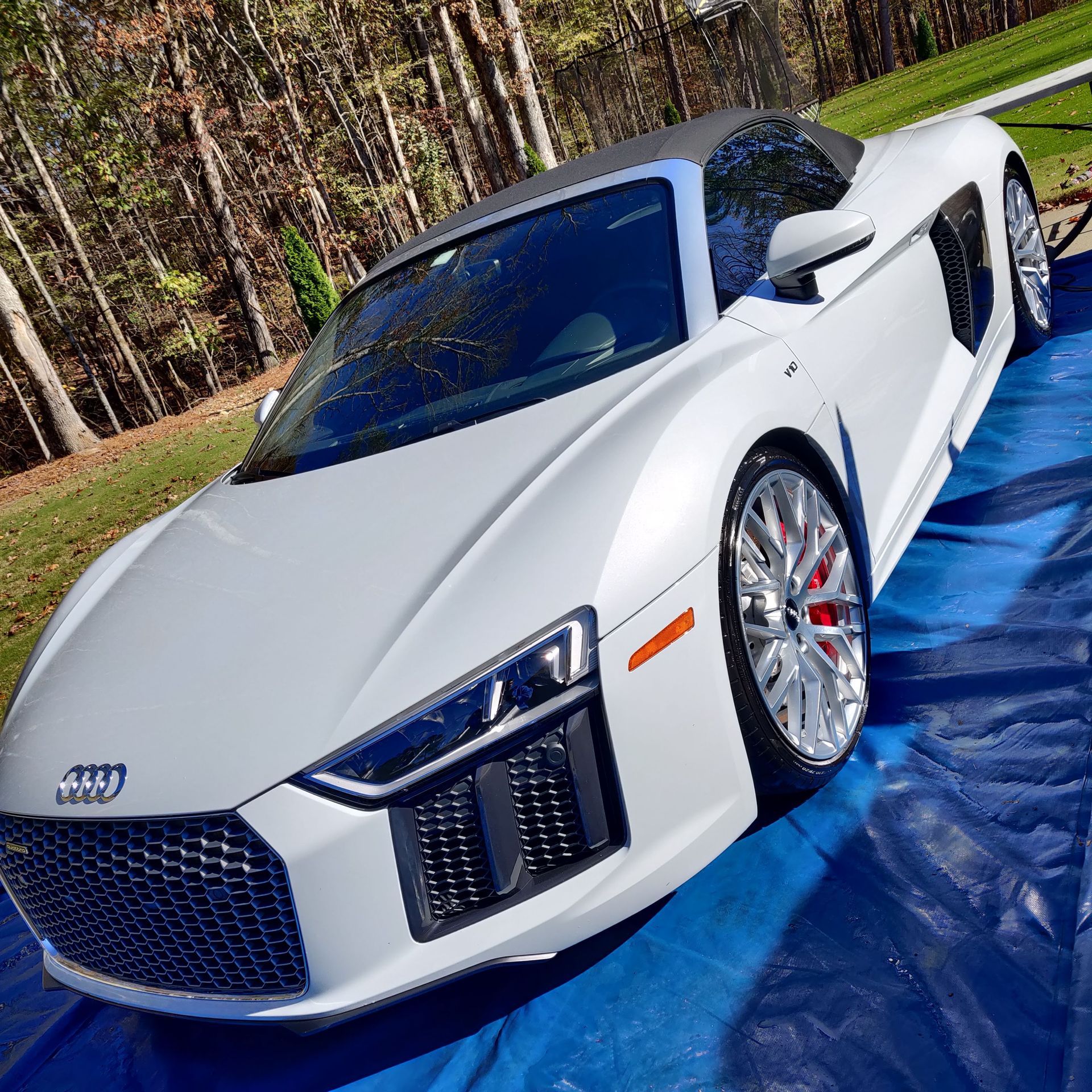White Audi R8 Spyder convertible on a blue tarp; chrome wheels with red calipers, parked outside, trees in the background.