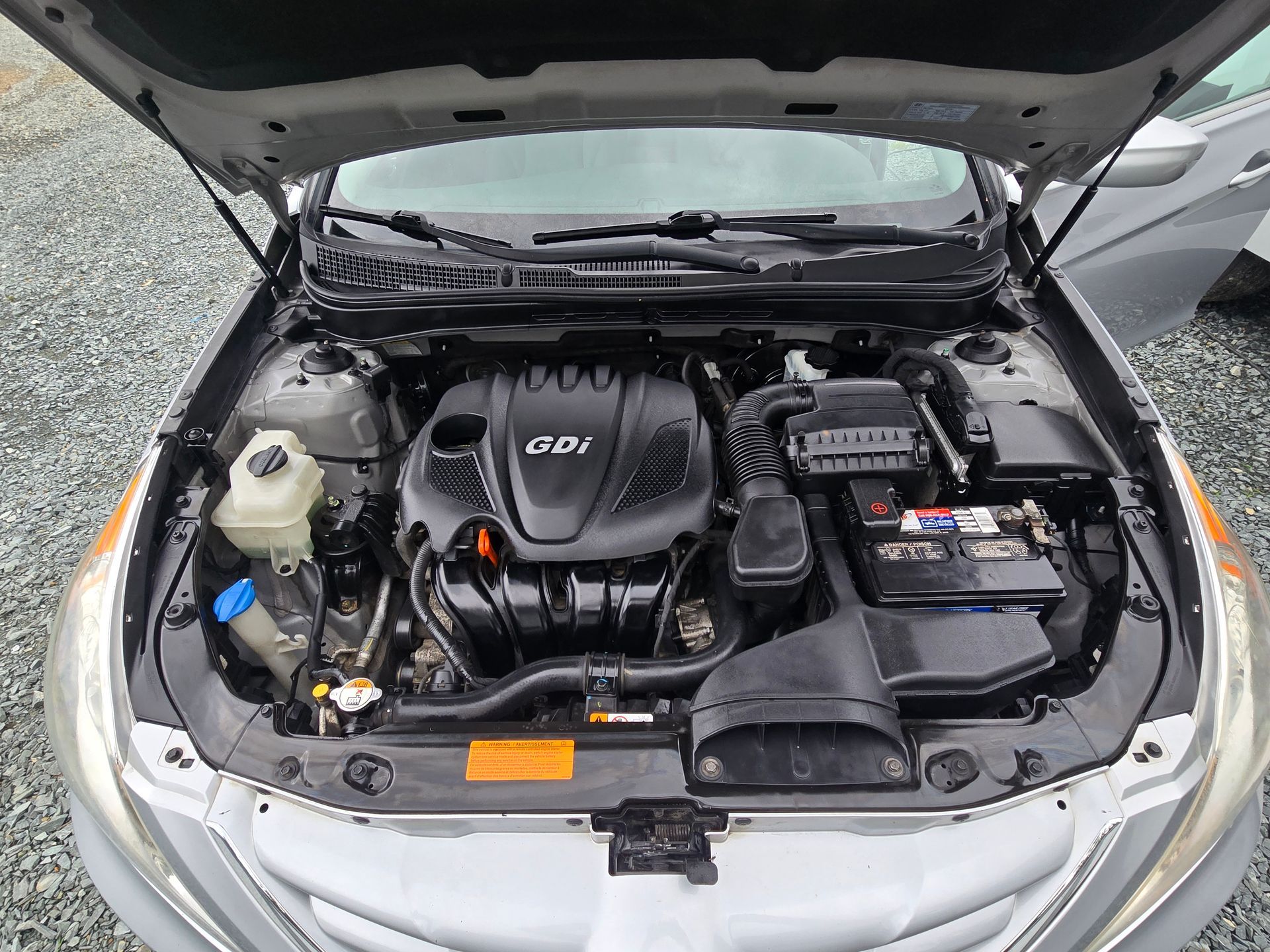 Car engine compartment with open hood, black engine cover, battery, and various components.