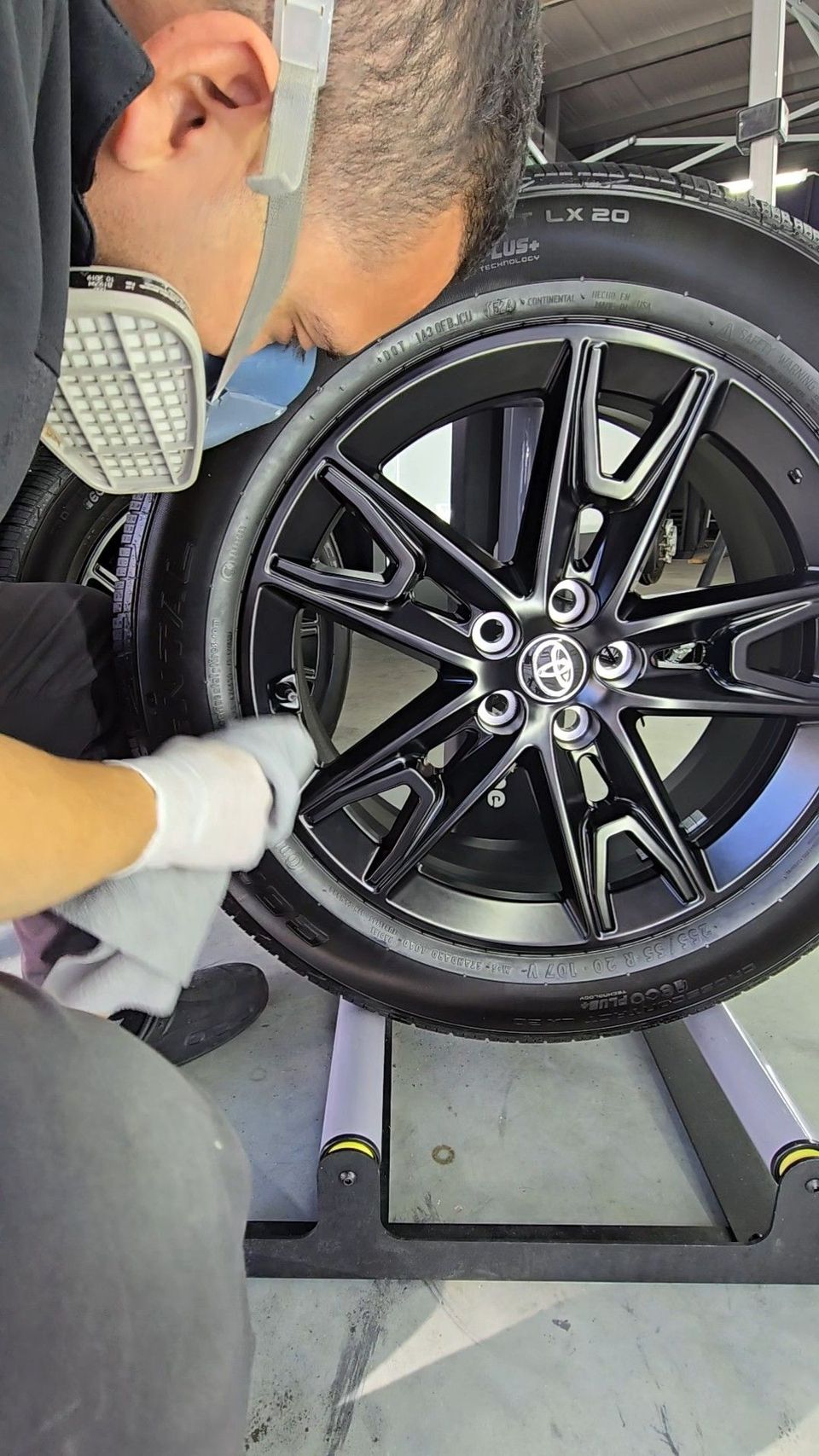 Person cleaning a black car rim with a cloth, wearing a respirator and gloves.