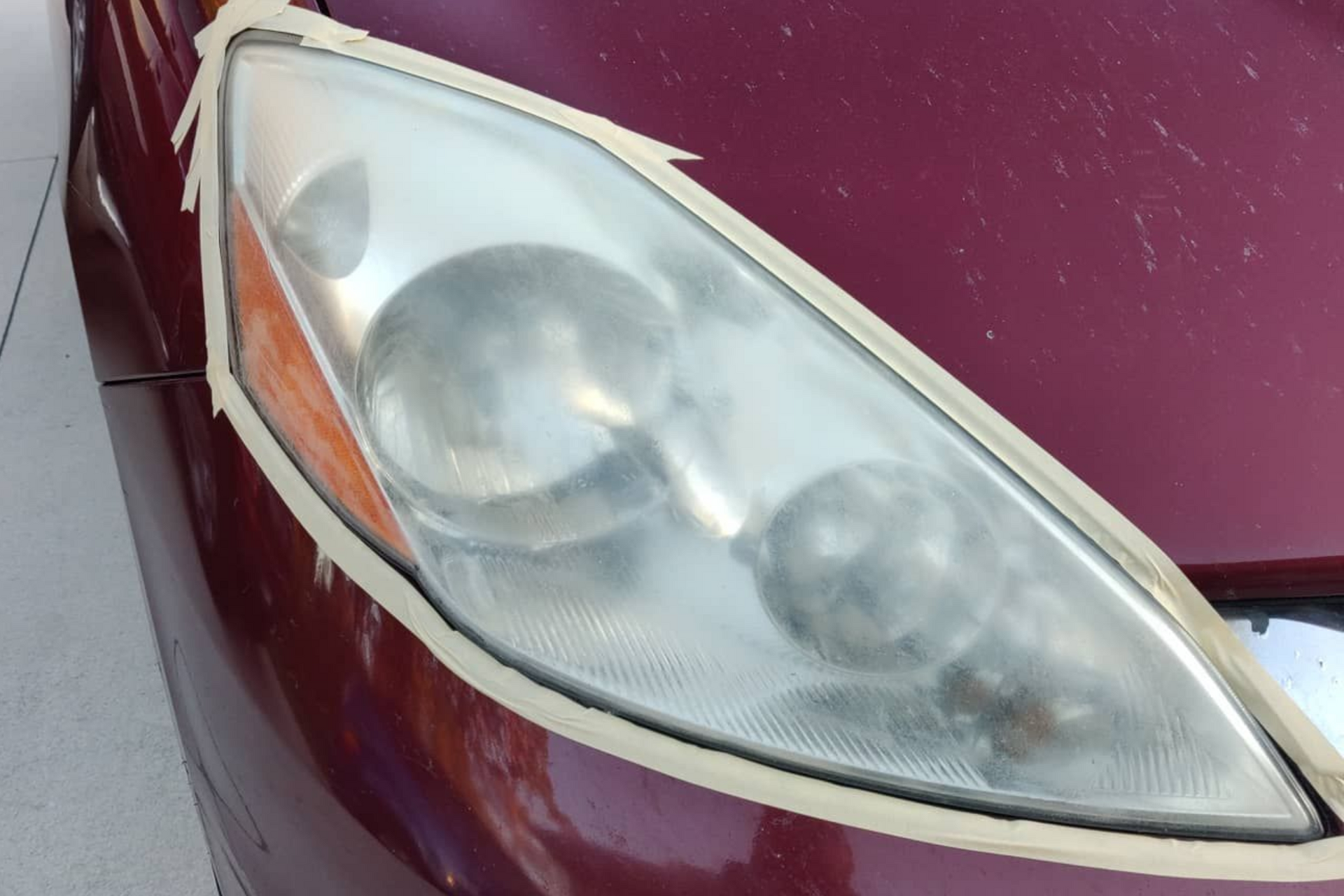 Close-up of a burgundy car's headlight, taped off, appearing hazy, ready for restoration.