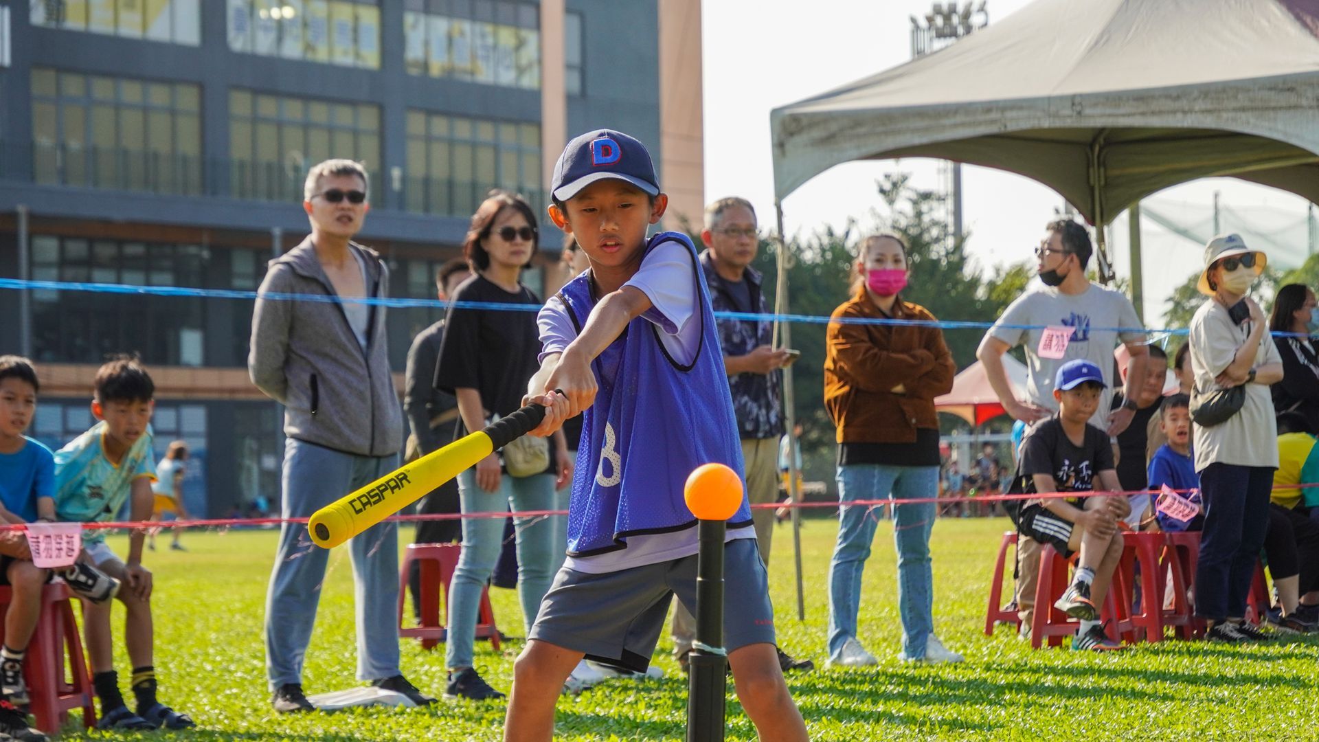 一個小男孩正在球上揮舞球棒，人們在一旁觀看。