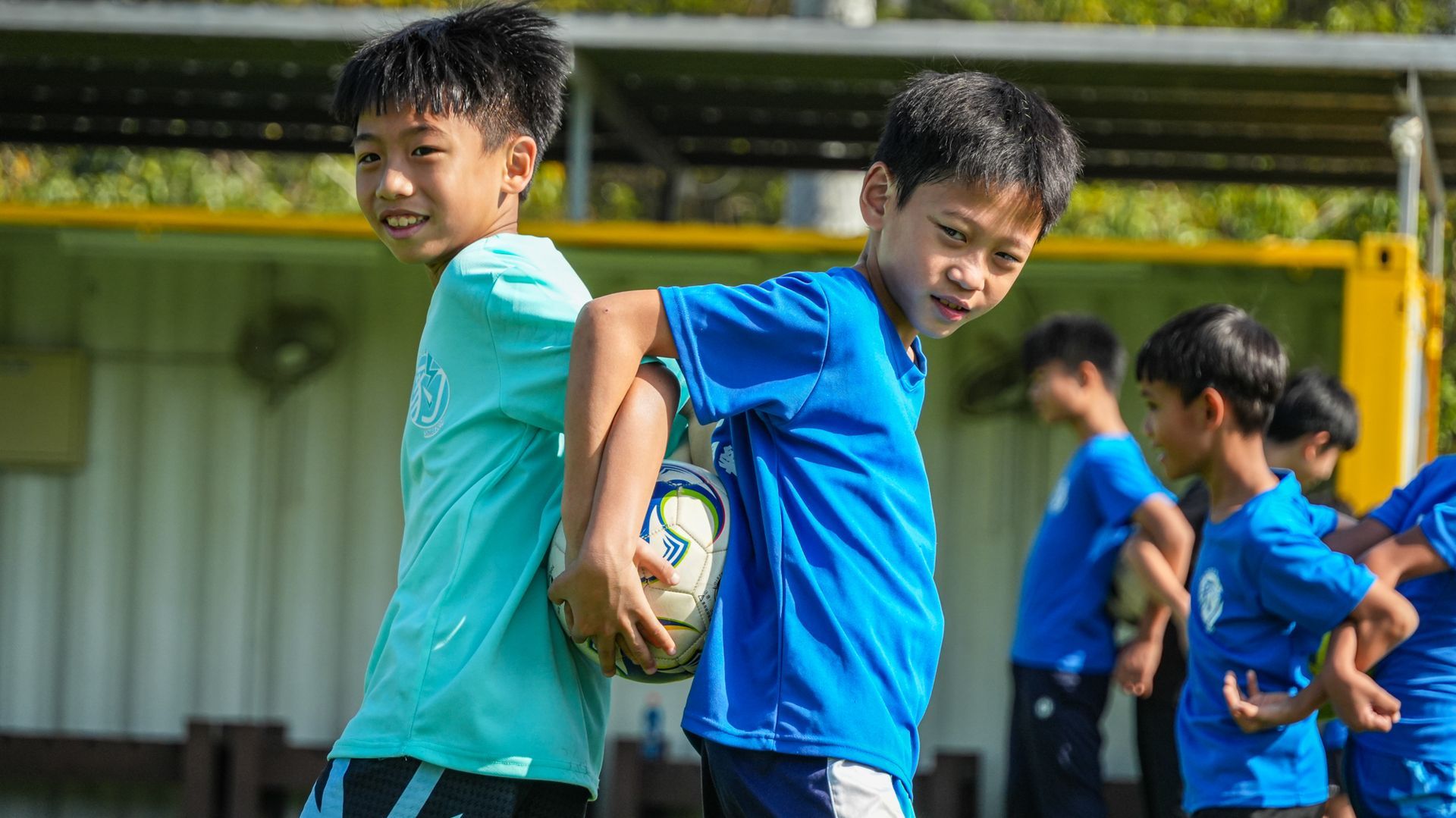 兩個小男孩背靠背站在足球場上，手裡拿著足球。