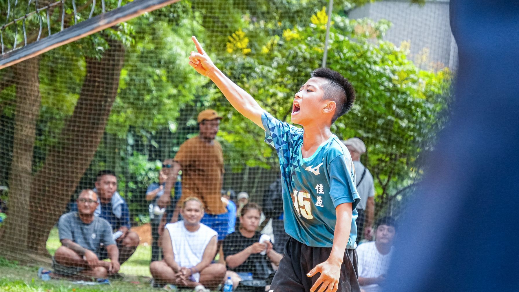 一個小男孩正在公園打排球，一群人在一旁觀看。