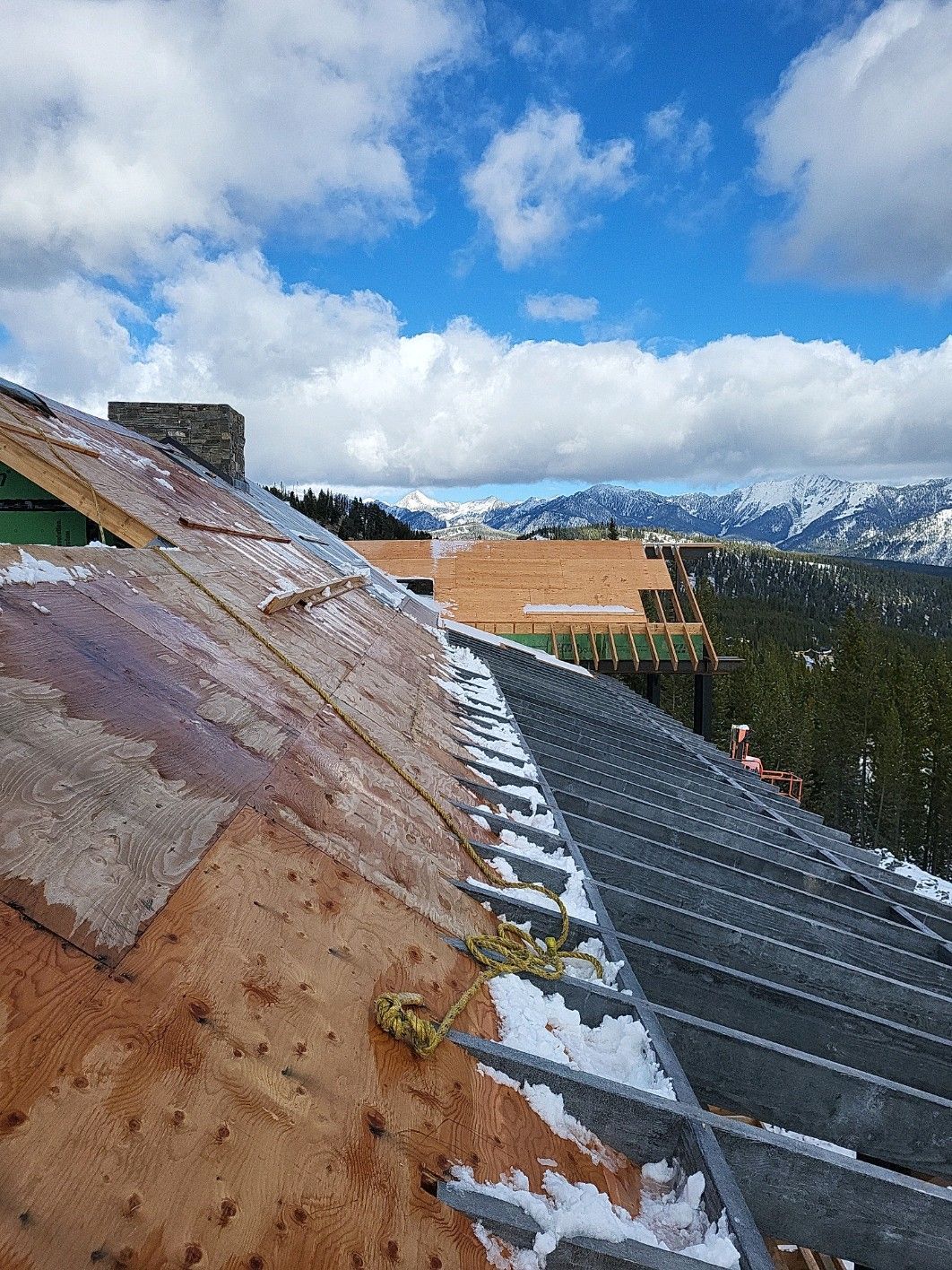 Roof under construction with mountain view, patches of snow, blue sky with clouds.