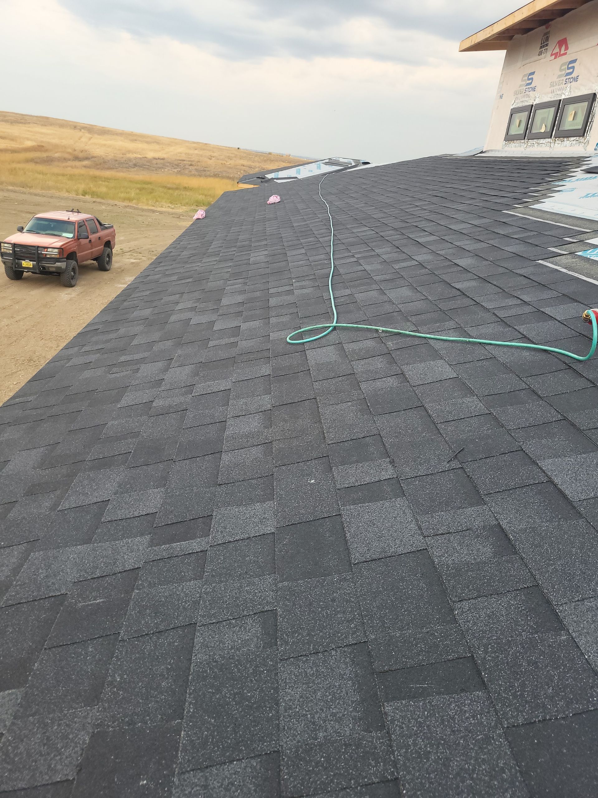 Roof shingles being installed on a house with a car and grassy hillside in background.