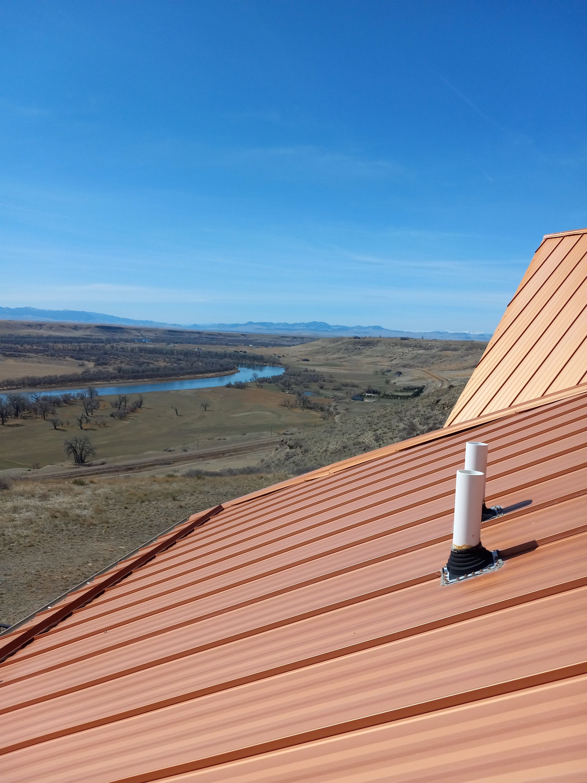 Copper metal roof with white pipe vent, overlooking a valley with a river under a blue sky.