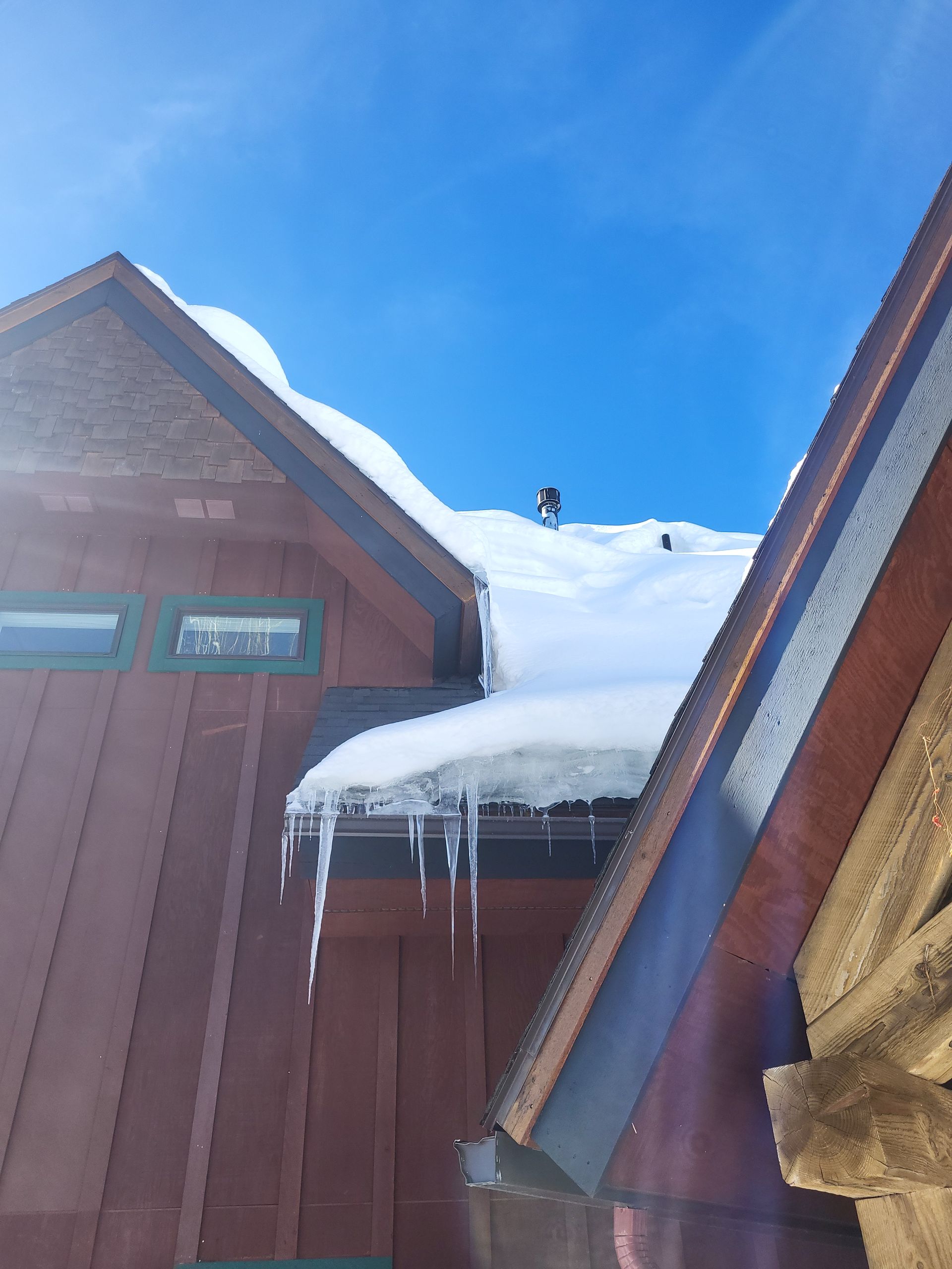 Snow-covered roof with icicles hangs over a red building against a bright blue sky.