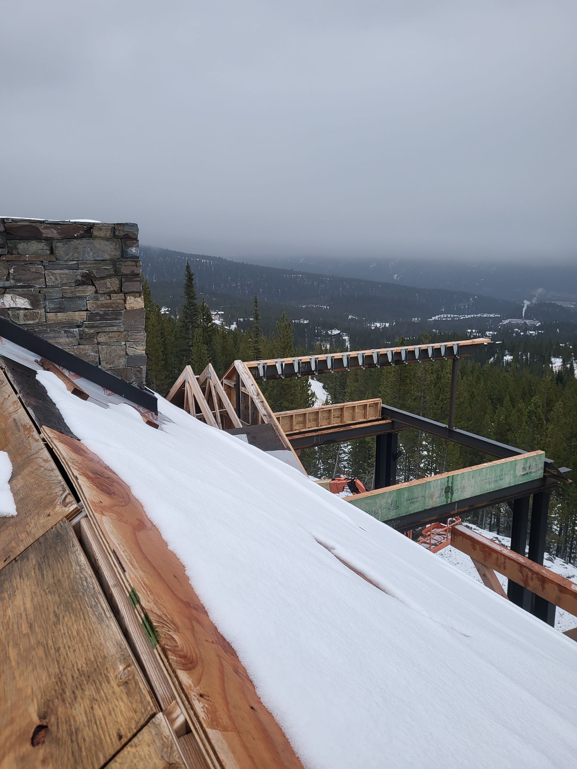 Rooftop under construction with snow, a stone chimney, and a distant view of trees and mountains under a cloudy sky.