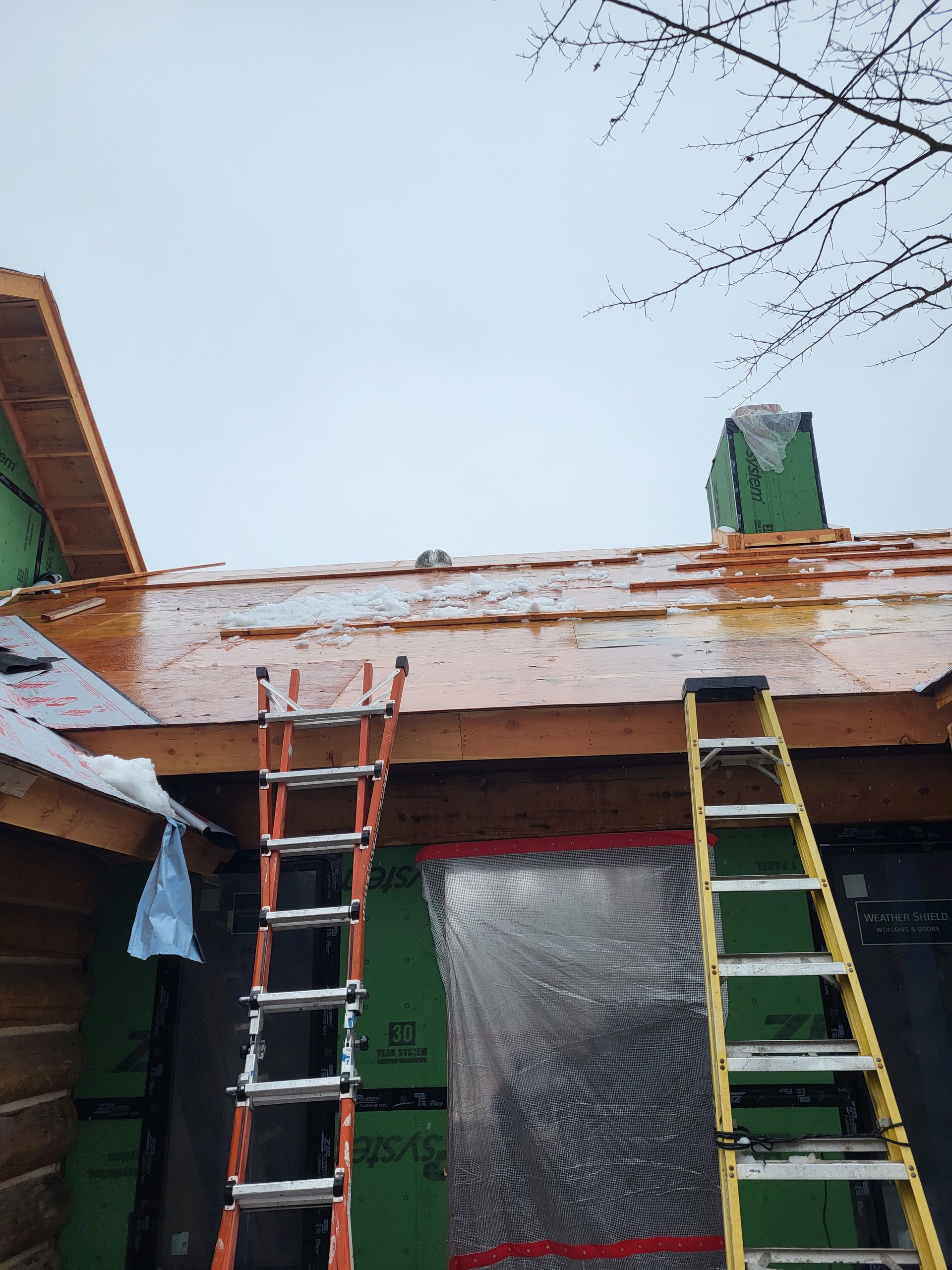Two ladders lean against a building under construction, roof partially covered with snow. Cloudy sky.
