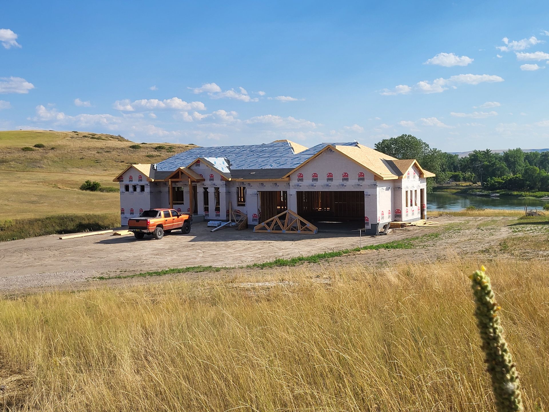 New house under construction, orange truck parked in front. Construction site on a grassy hill, with a pond visible.