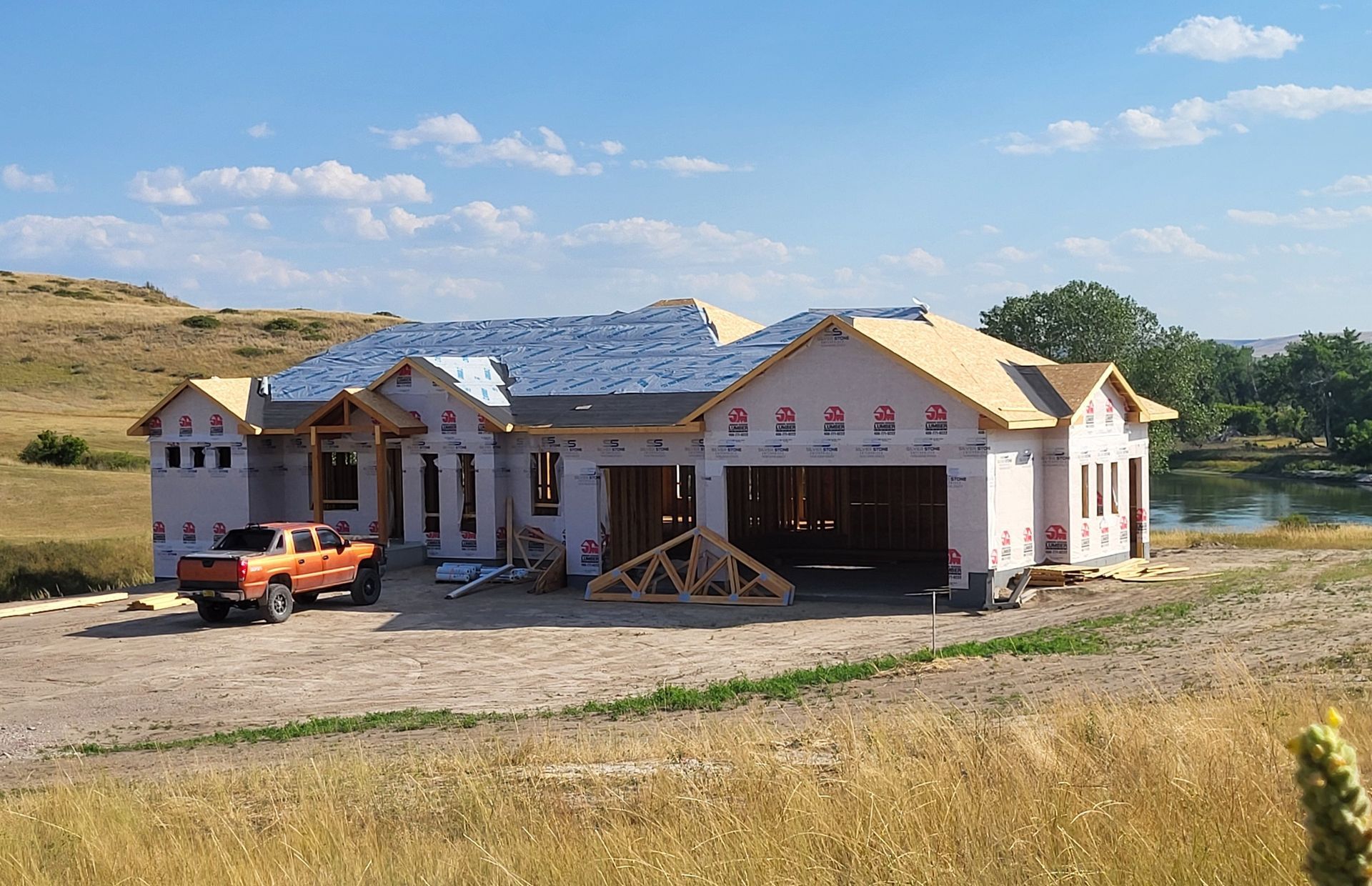 House under construction with orange truck, surrounded by grassy field and a pond.