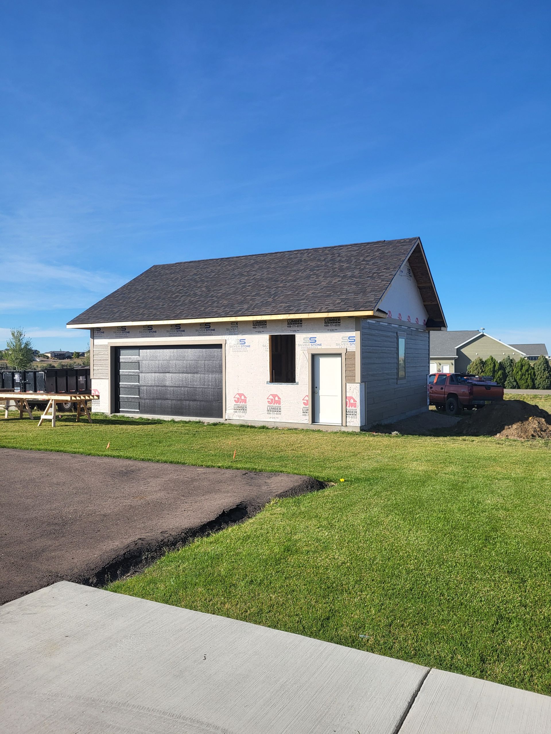 Unfinished gray building with a dark roof and garage door. Construction is in progress on a green lawn.